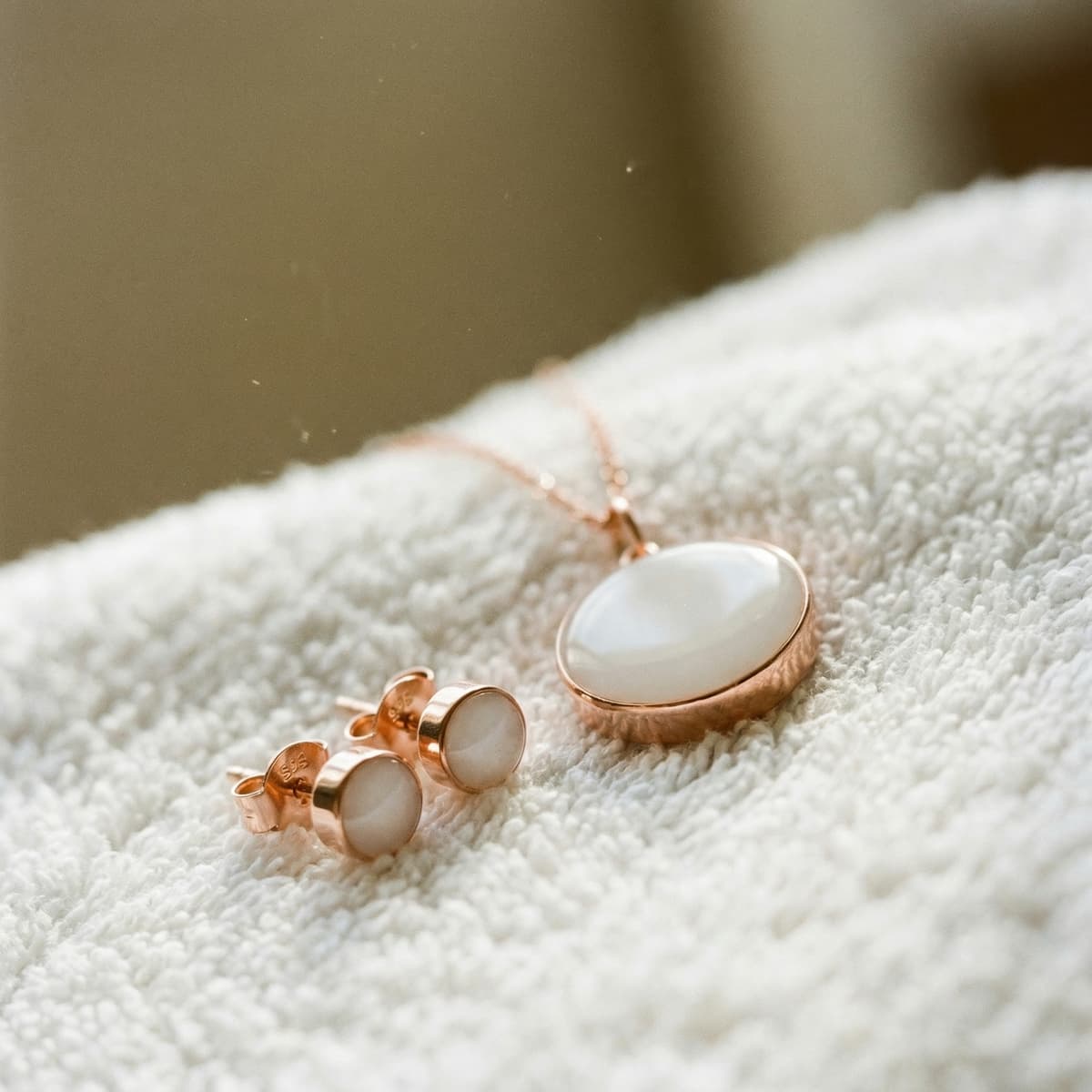 Rose gold breastmilk jewelry set with oval pendant necklace and matching stud earrings featuring milky white resin stones, celebrating a 1 year breastfeeding award, photographed on a soft white towel in warm warm light.