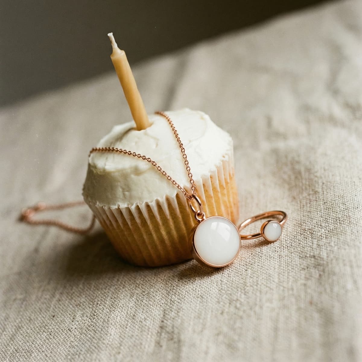 Cupcake with white frosting and a single candle beside rose-gold breastmilk jewelry pendant and ring, styled as a 1 year breastfeeding award keepsake.