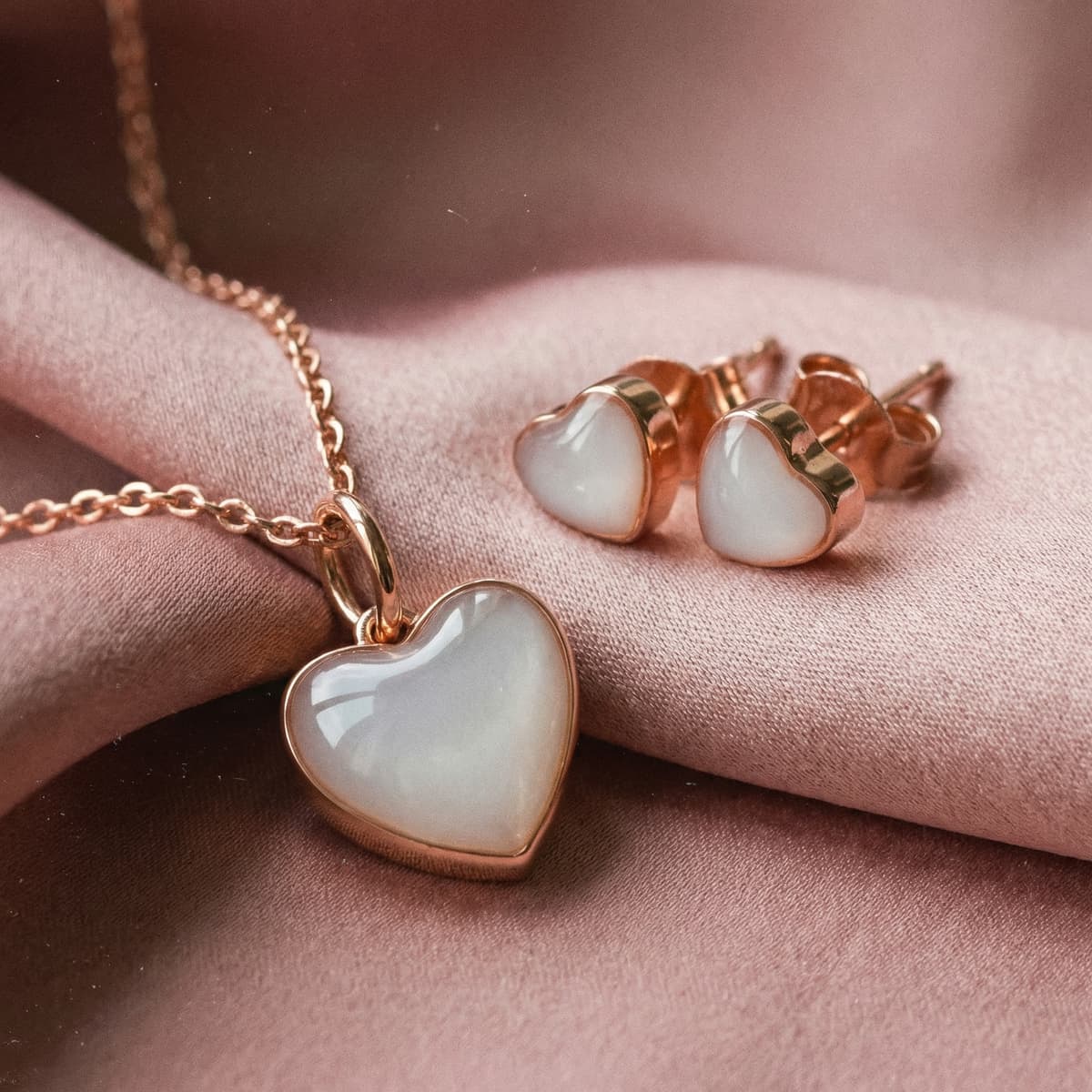 Rose-gold heart pendant necklace with matching heart stud earrings filled with creamy white breastmilk resin, displayed on soft pink fabric as a sentimental 1 year breastfeeding gift.
