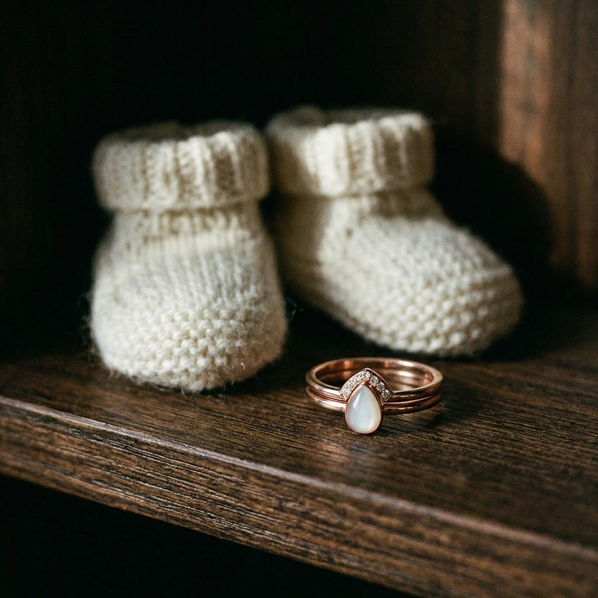 Delicate breastmilk jewelry ring with a pear-shaped milky resin stone displayed beside tiny knitted baby booties on a wooden shelf, a sentimental baby keepsake for mom.