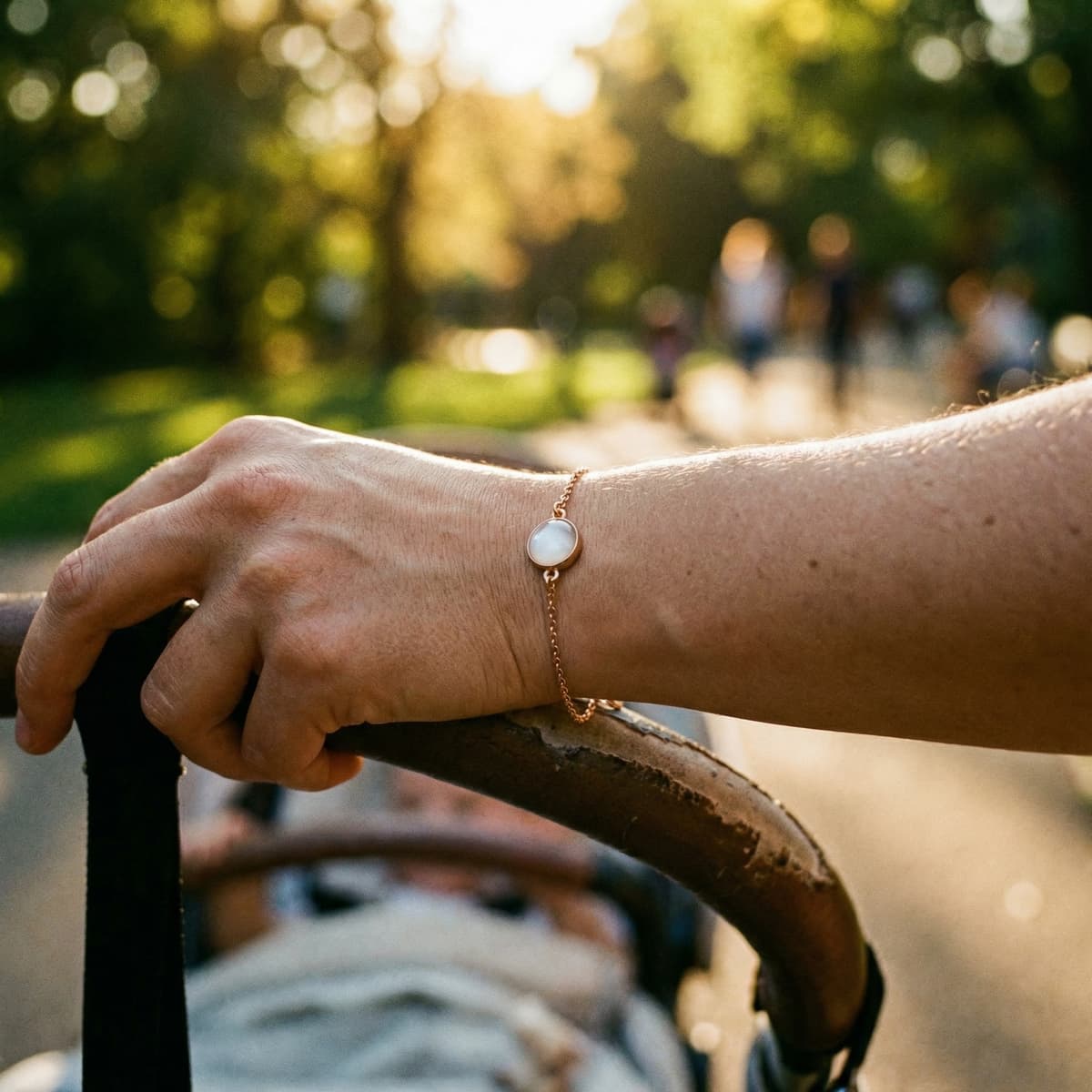 Close-up outdoors of a mother’s hand holding a stroller handle, wearing a delicate gold bracelet with a milky-white stone, a sentimental baby keepsake for mom.