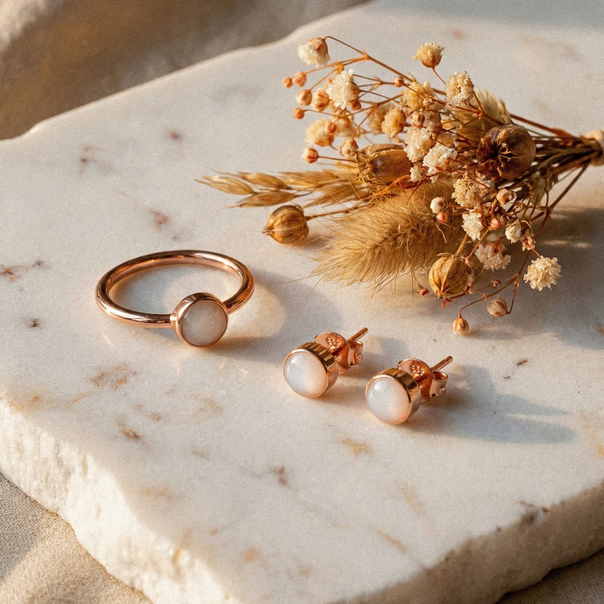 Elegant rose-gold breastmilk keepsake ring and matching stud earrings with milky-white resin stones on a marble slab beside dried flowers, perfect as one of the best push presents for new moms.