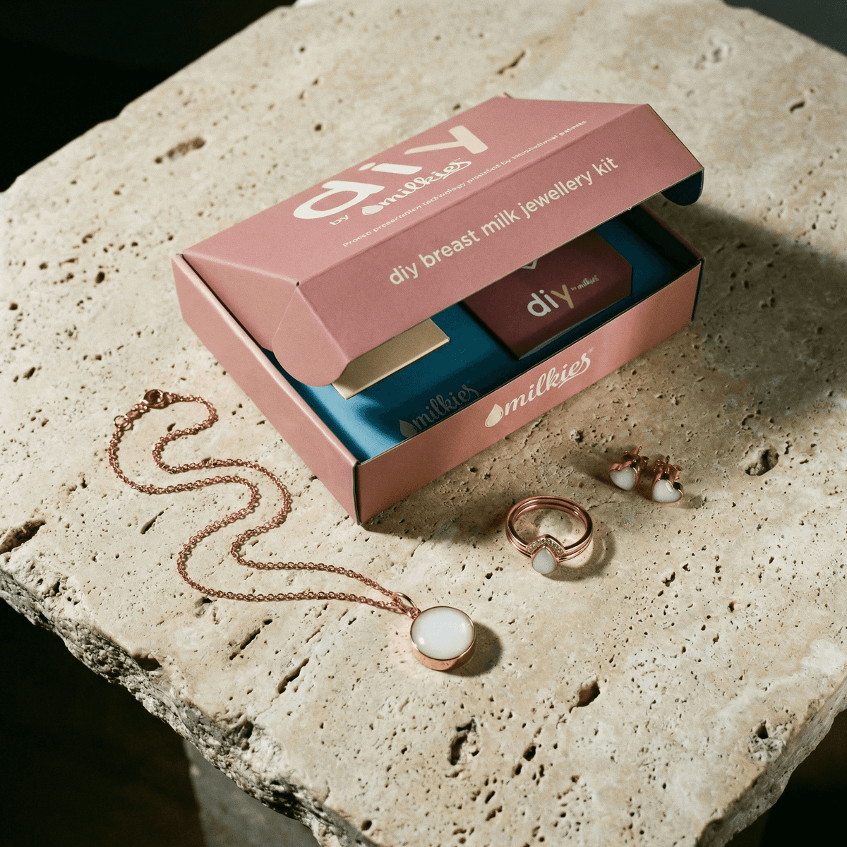 Open DIY by MILKIES breast milk jewellery diy kit box on a stone surface with rose gold pendant necklace, ring, and matching stud earrings featuring milky white resin stones.
