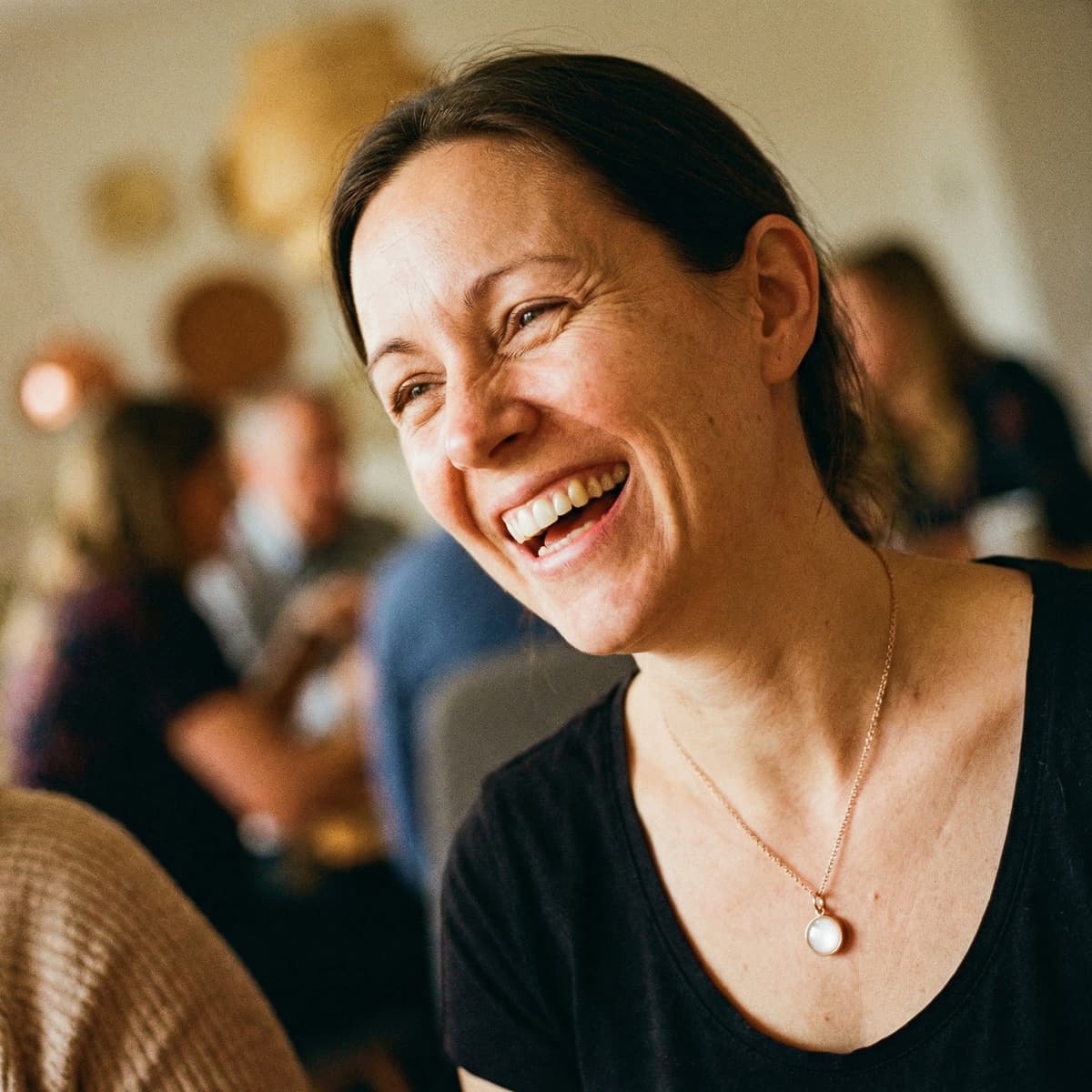 Smiling mother wearing a breastmilk keepsake necklace with a milky-white pendant, capturing a warm breastfeeding celebration in a cozy indoor setting.