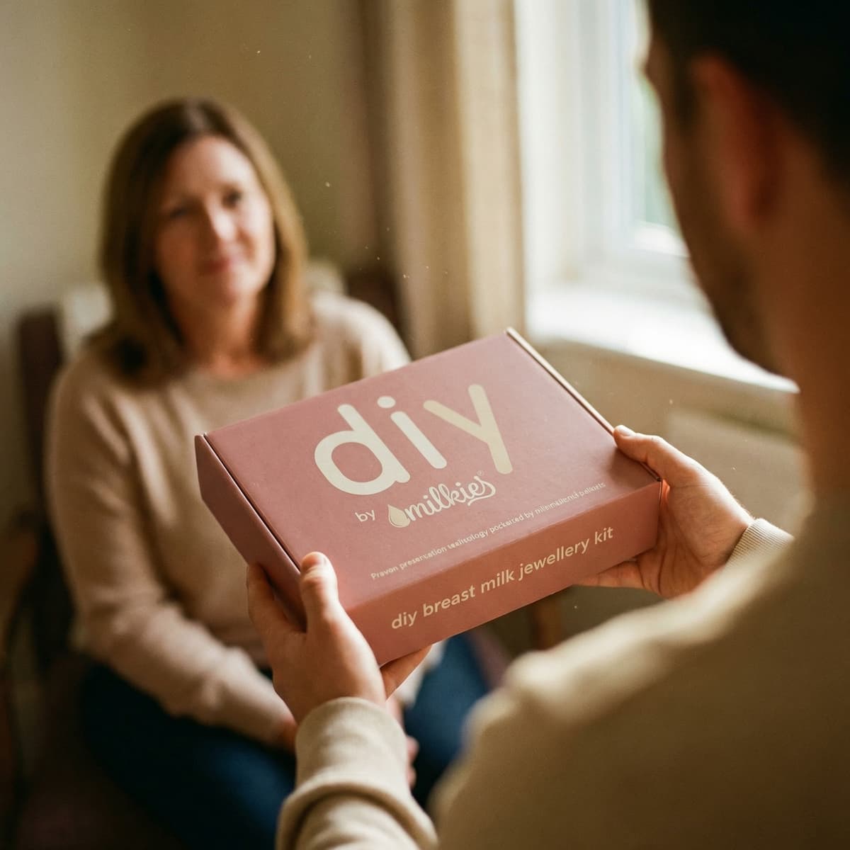 Woman seated as another person hands her a pink DIY by MILKIES breast milk jewellery kit box, shown as a thoughtful breastfeeding gift for creating a keepsake at home.