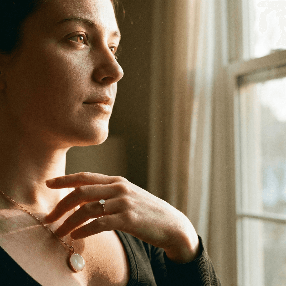 Woman gazes out a window in warm natural light, wearing a breastmilk pendant necklace and ring as a sentimental breastfeeding journey gift.