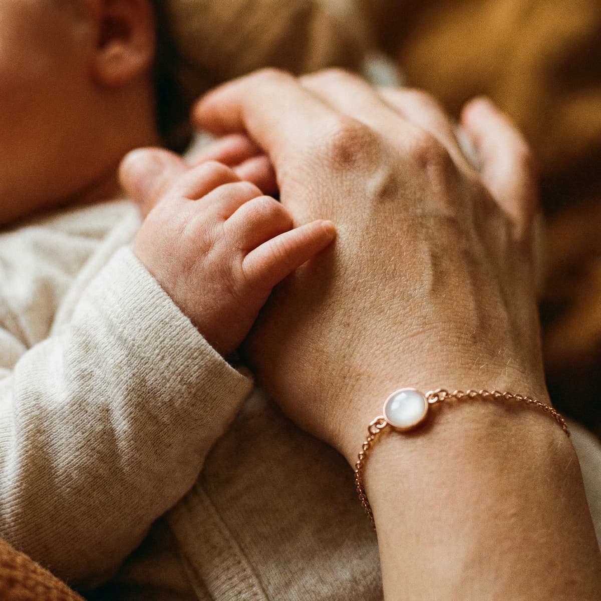 Mother holding baby’s hand while wearing a delicate rose-gold breastmilk bracelet featuring a milky white resin stone keepsake