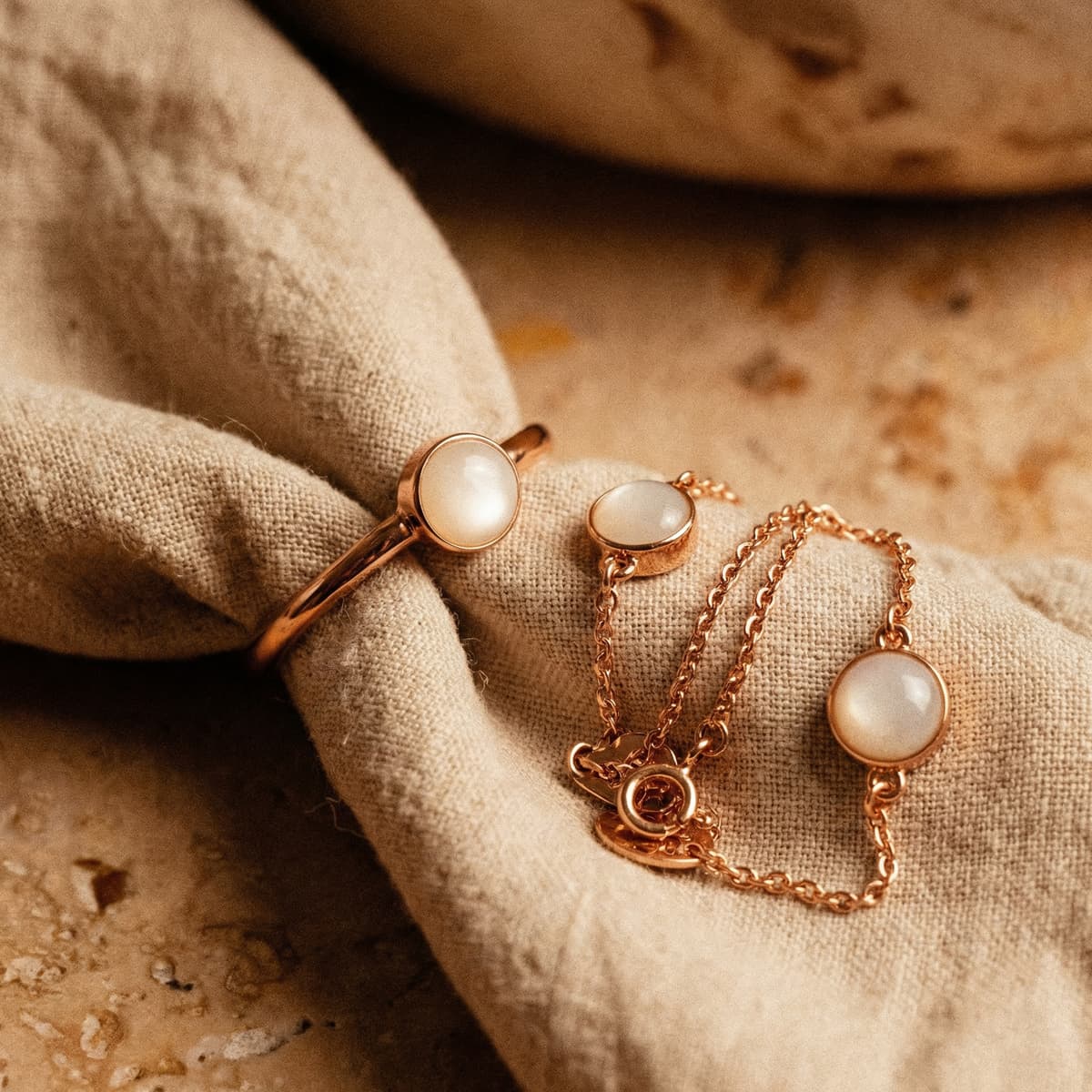 Rose gold keepsake jewelry set featuring a breastmilk bracelet with a round milky-white resin charm, paired with a matching ring and stud earrings on beige linen fabric.