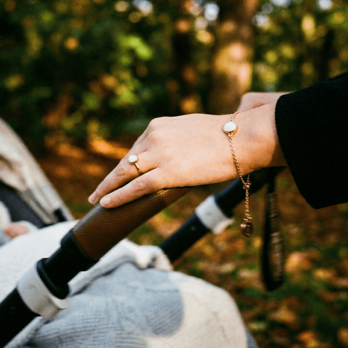Mother’s hand holding a stroller handle, wearing a delicate gold bracelet with a milky-white stone made using a breastmilk bracelet diy kit.