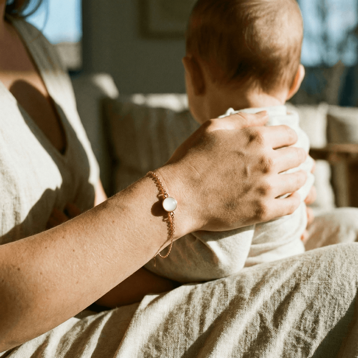 Mother holding her baby in warm light, wearing a delicate keepsake bracelet with a milky stone—showing why a breastmilk bracelet diy kit is a meaningful at-home way to preserve motherhood memories.