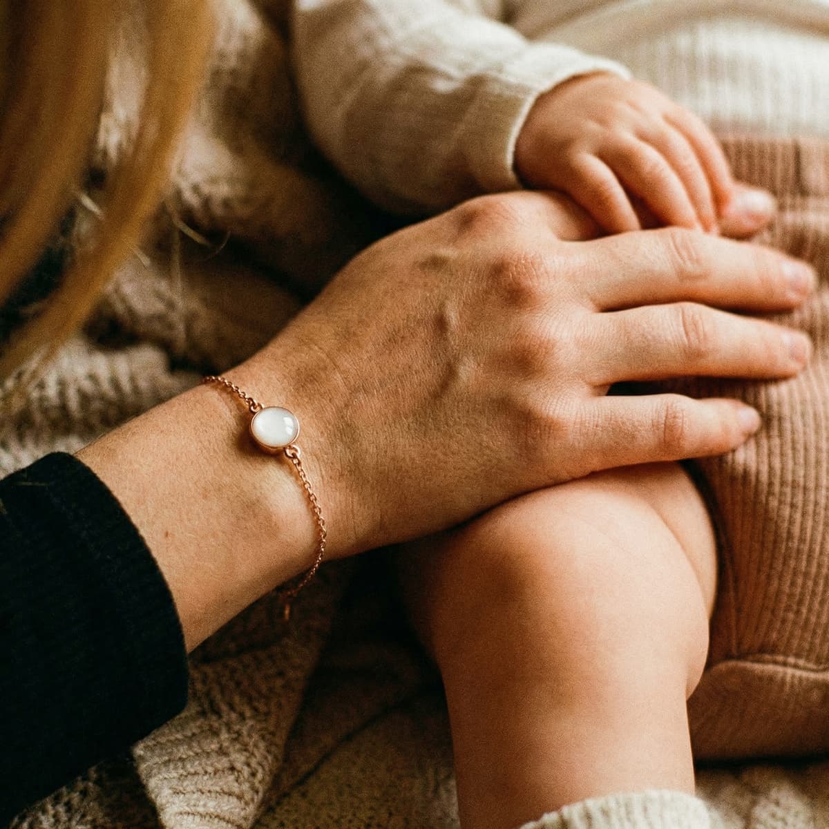 Mother holding a baby’s hand while wearing a delicate rose-gold keepsake bracelet with a milky white resin stone made using a breastmilk bracelet kit.