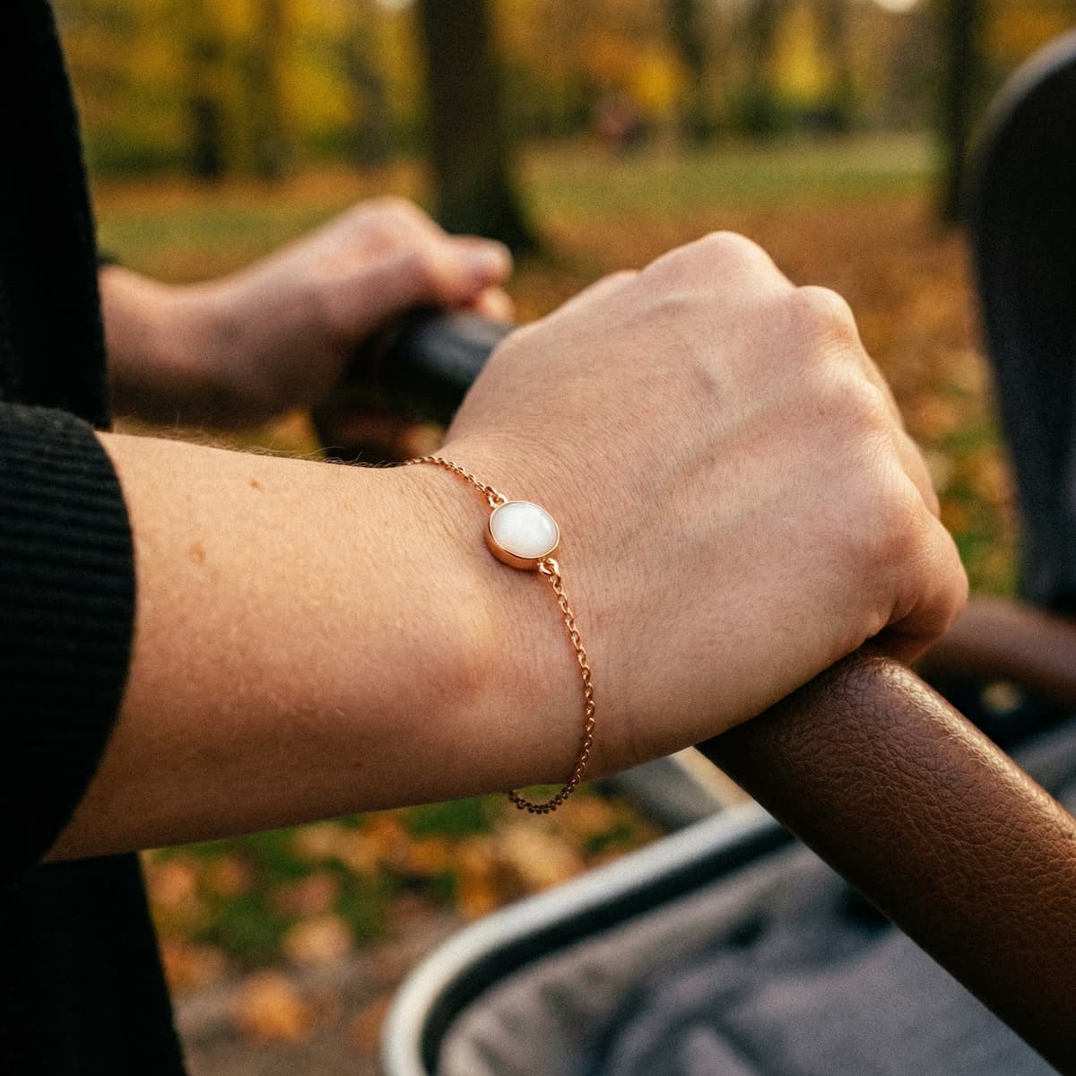 Hand wearing a delicate rose-gold chain keepsake bracelet with a round milky-white resin stone made with a breastmilk bracelet kit, pushing a stroller outdoors in autumn.