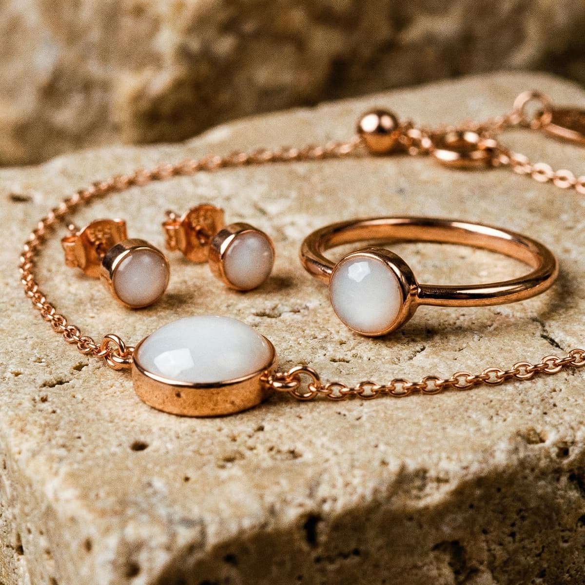 Rose gold jewelry set on textured stone background featuring a delicate chain bracelet from a breastmilk bracelet kit with milky white resin stone, matching stud earrings, and a minimalist ring