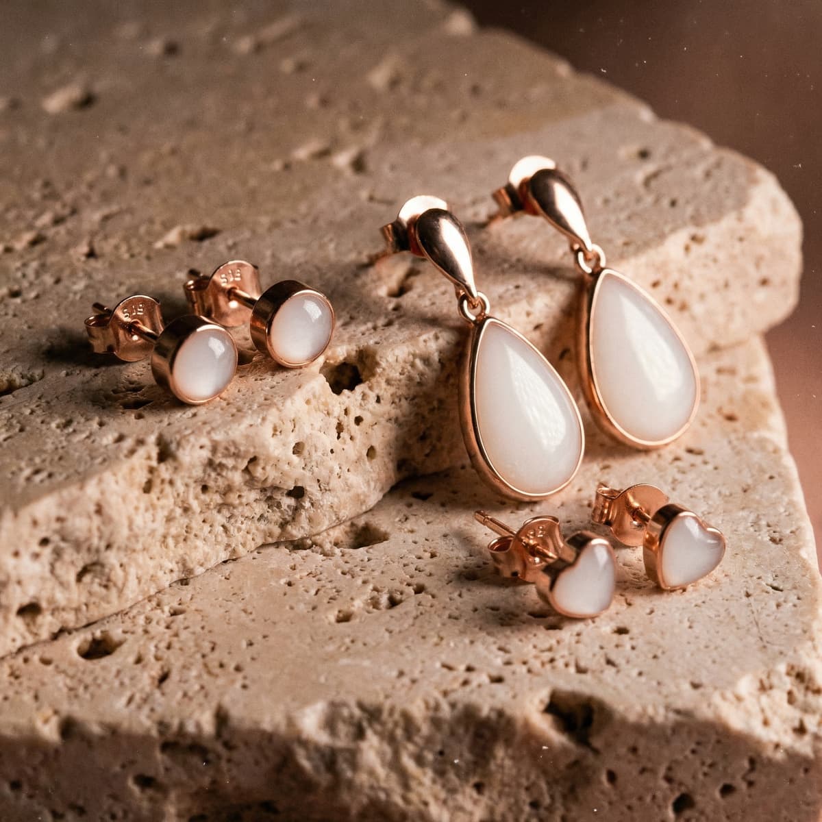 Rose gold jewelry set on textured stone featuring a breastmilk earring collection with milky white resin stud earrings, heart-shaped studs, and teardrop dangle earrings