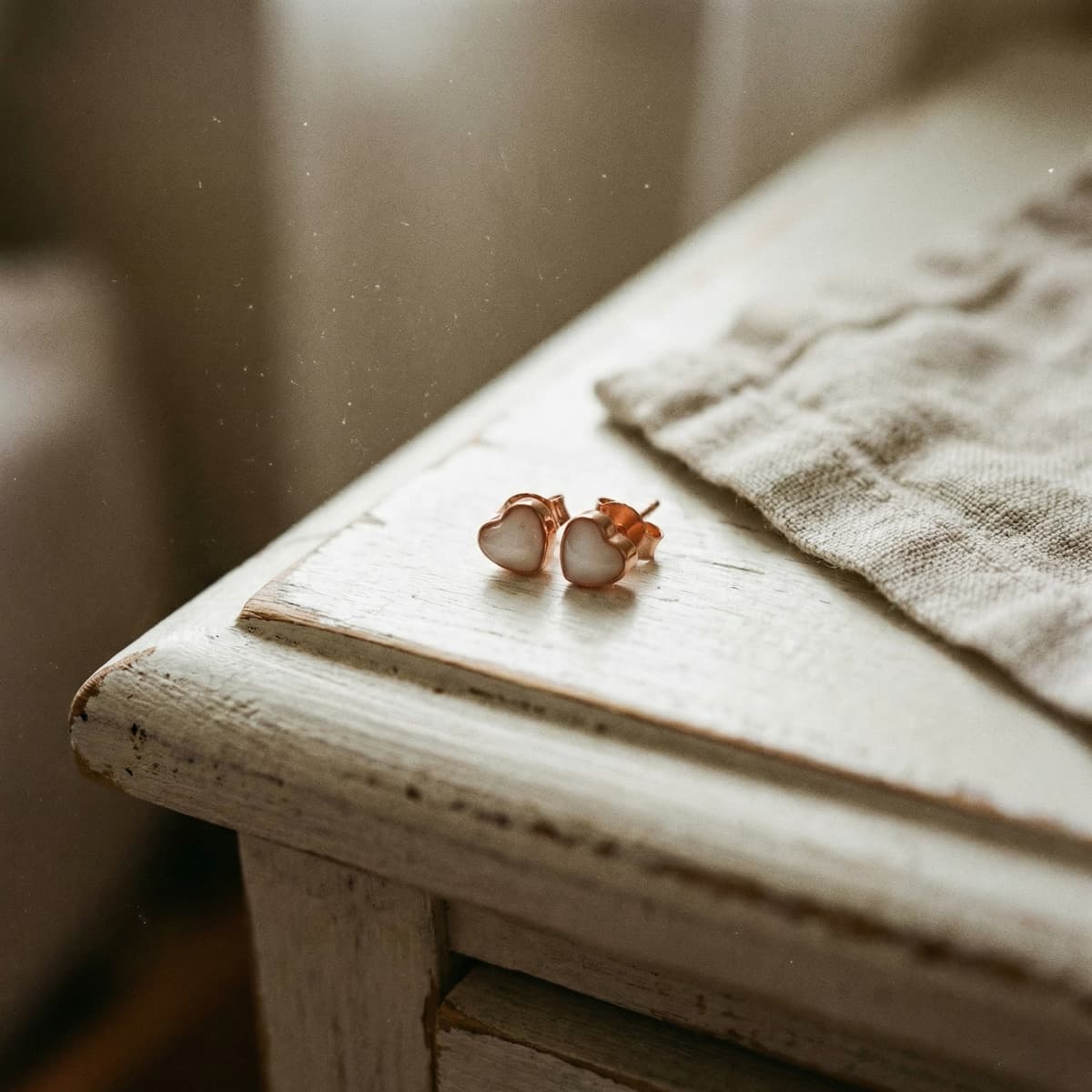 Rose-gold heart stud earrings made with a breastmilk earring diy kit, displayed on a white wooden table beside a soft linen cloth in warm natural light.