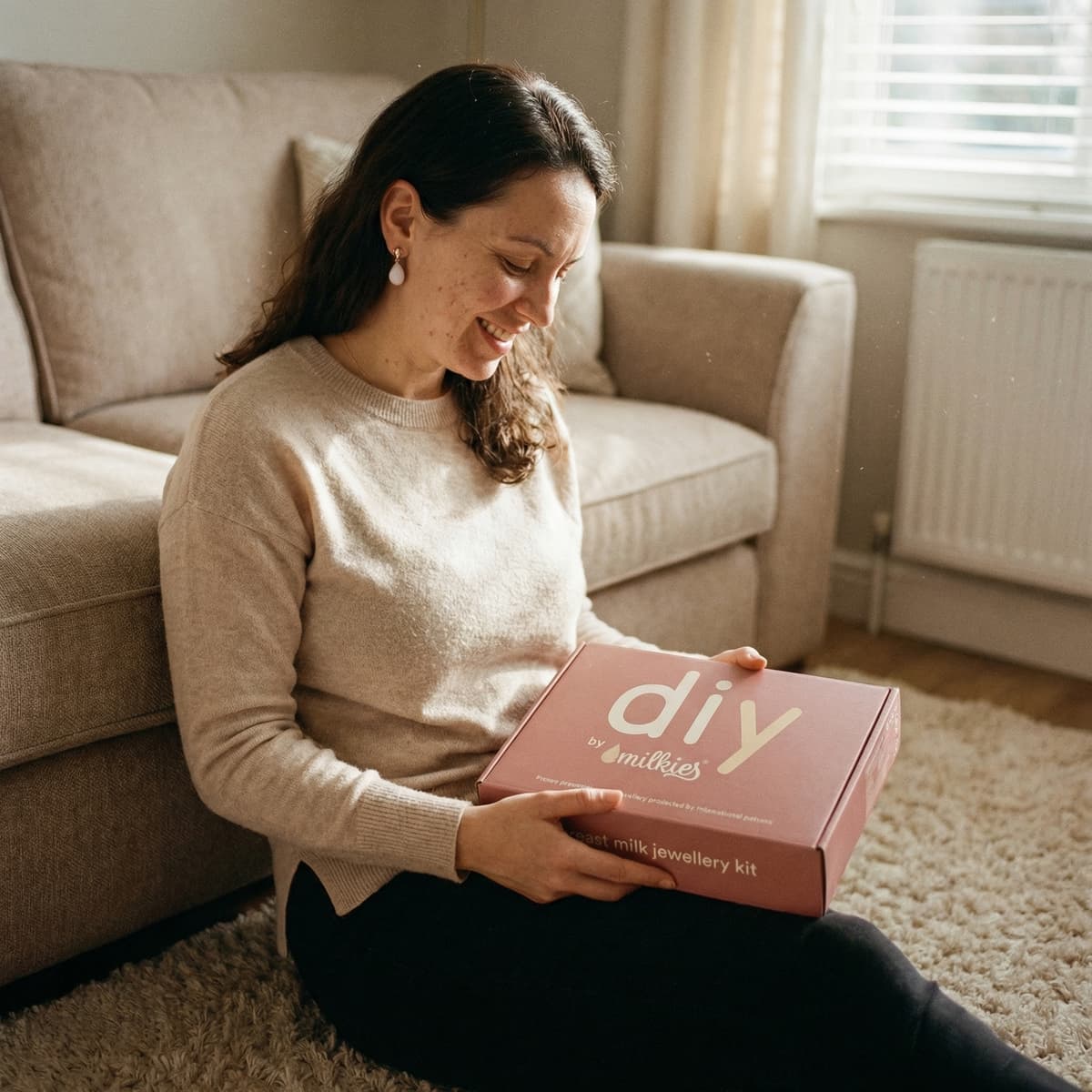 Smiling mother at home holding a DIY by MILKIES box, showing why a breastmilk earring diy kit is a convenient, private way to create a hands-on keepsake at home.