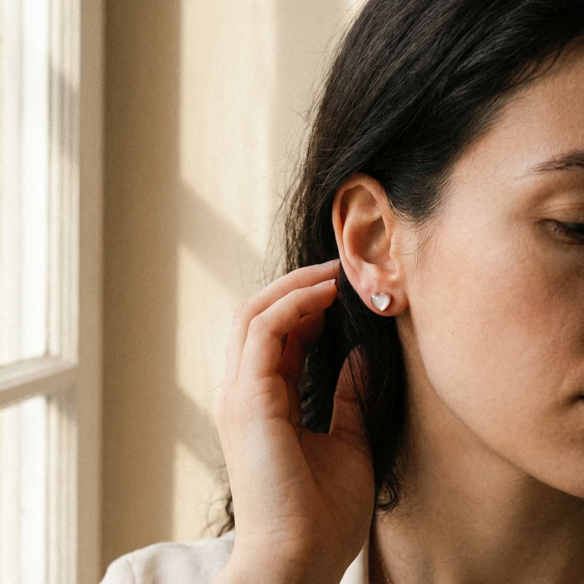 Close-up of a woman wearing small heart-shaped breastmilk keepsake stud earrings in soft window light, showcasing the finished jewelry made with a breastmilk earring kit.
