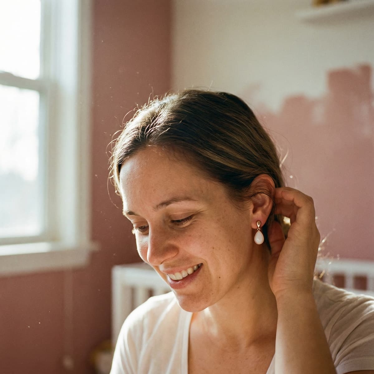Smiling mother in a sunlit room wearing white teardrop earrings, showing why a breastmilk earring making kit is a meaningful DIY way to create a personal keepsake at home.
