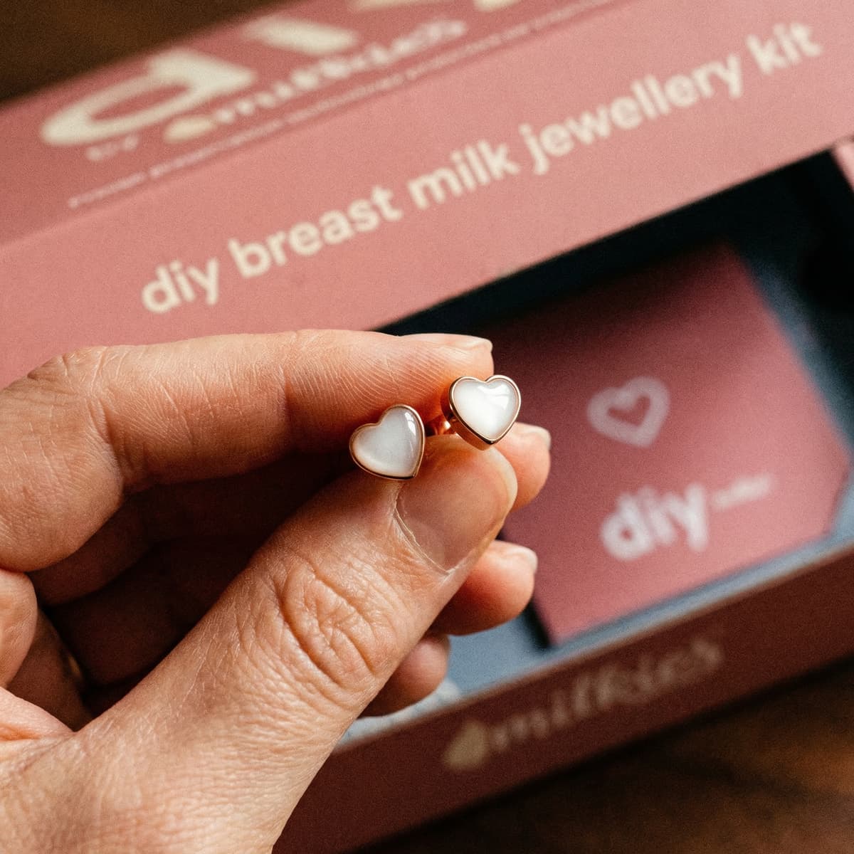 Hand holding glossy heart-shaped breastmilk earrings in gold settings, shown in front of a DIY by MILKIES breast milk jewelry kit box.