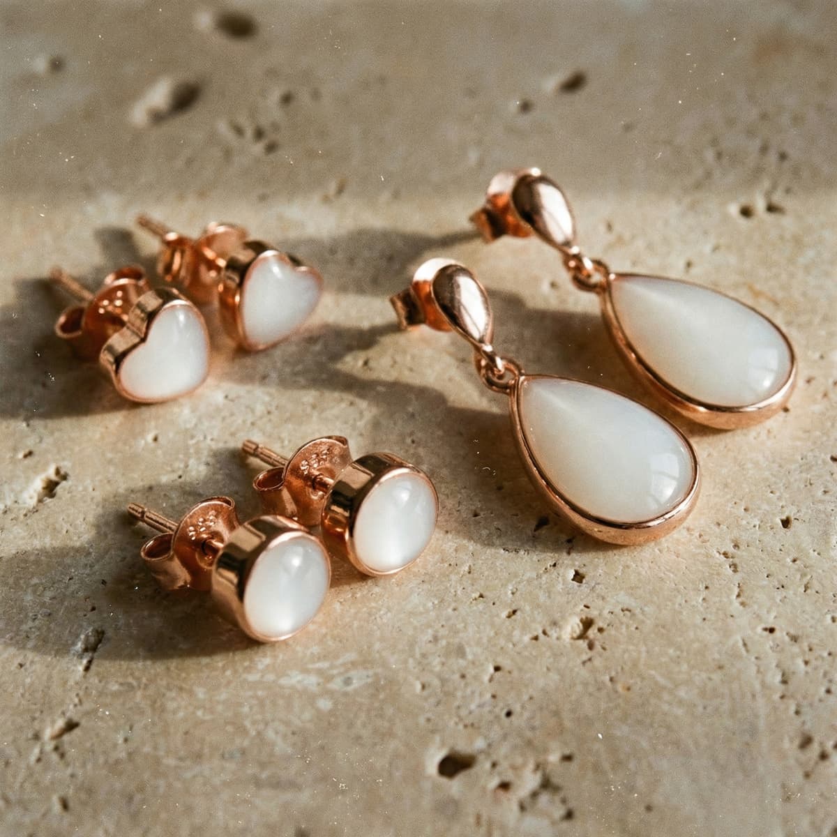 Rose gold breastmilk earrings set featuring milky white resin stones, with teardrop dangle, round stud, and heart stud designs arranged on a textured stone background.
