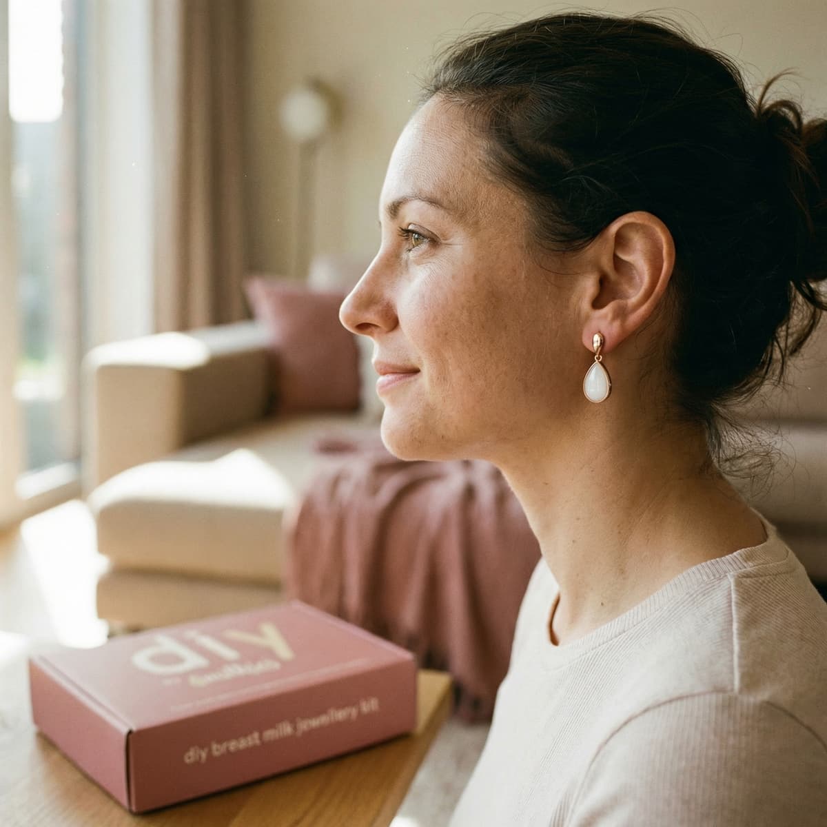 Woman at home wearing elegant breastmilk earrings beside a DIY by MILKIES breast milk jewelry kit box, highlighting why making a private, meaningful keepsake yourself is a great idea.