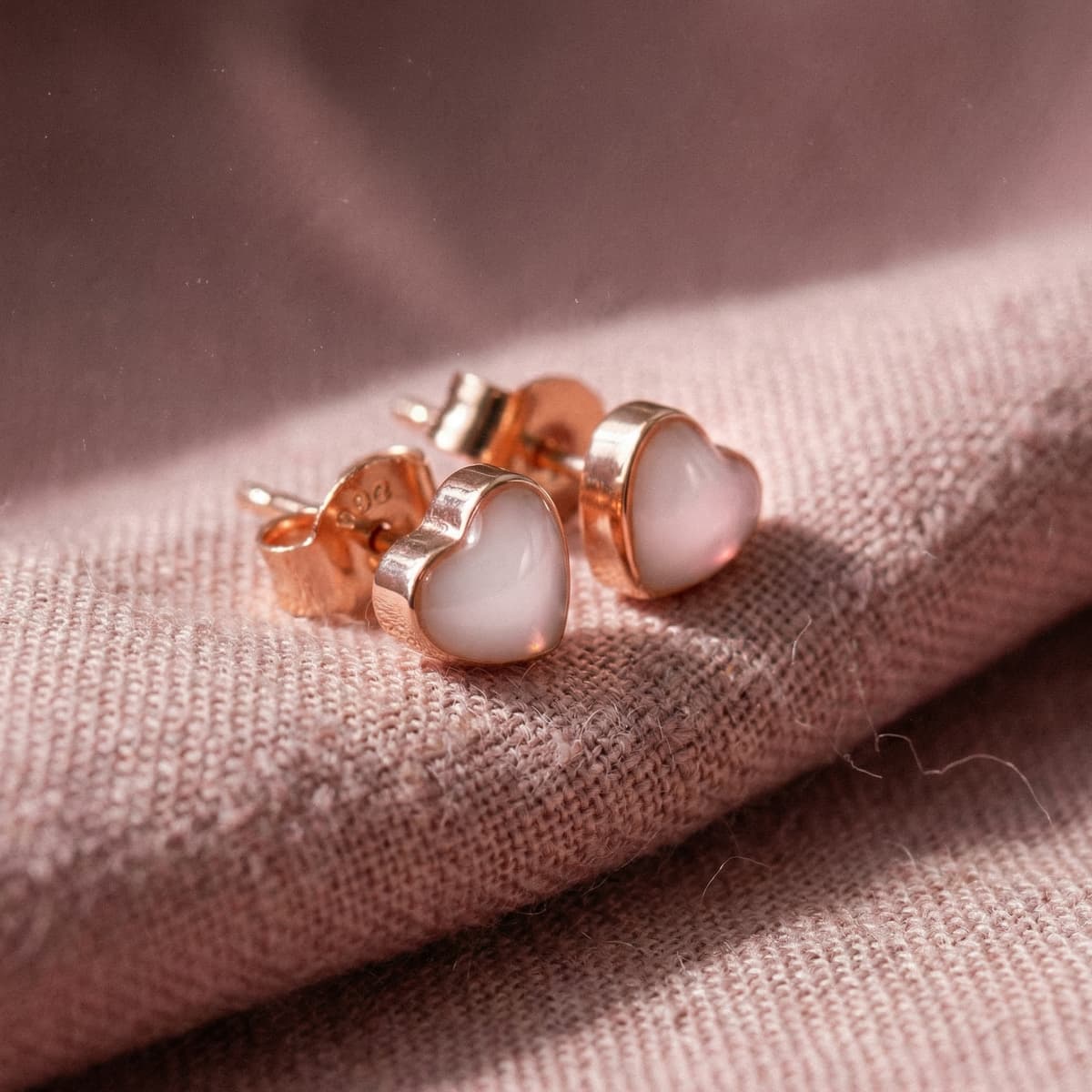 Heart-shaped rose gold stud earrings with milky white resin stones, made with a breastmilk earrings kit keepsake jewelry design on soft blush fabric.
