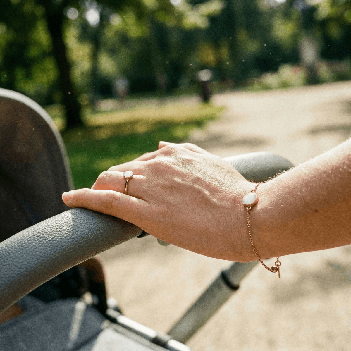 Mother’s hand pushing a stroller in soft sunlight, wearing minimalist gold breastmilk jewellery ring and matching bracelet with milky white resin stones outdoors.