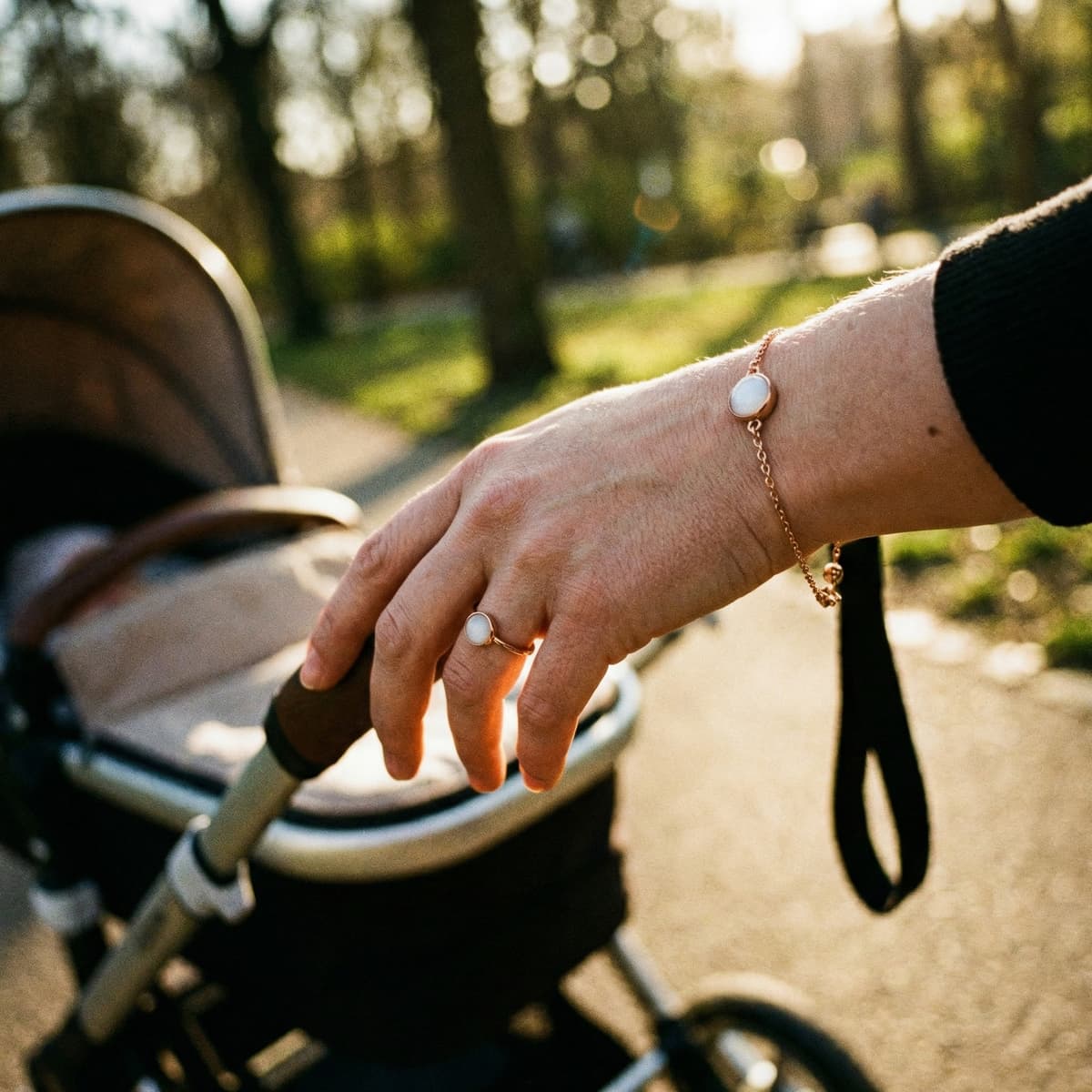 Mother’s hand pushing a baby stroller outdoors in warm sunlight, wearing a delicate gold bracelet and ring with milky-white stones as breastmilk keepsake jewelry.
