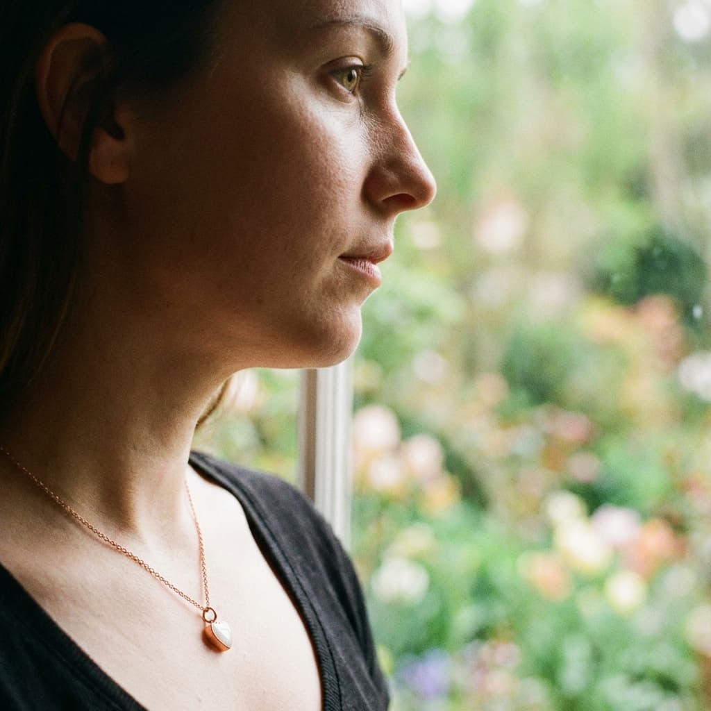 Mother in side profile wearing a delicate gold pendant made with a breastmilk necklace diy kit, standing by a window with a soft garden background.