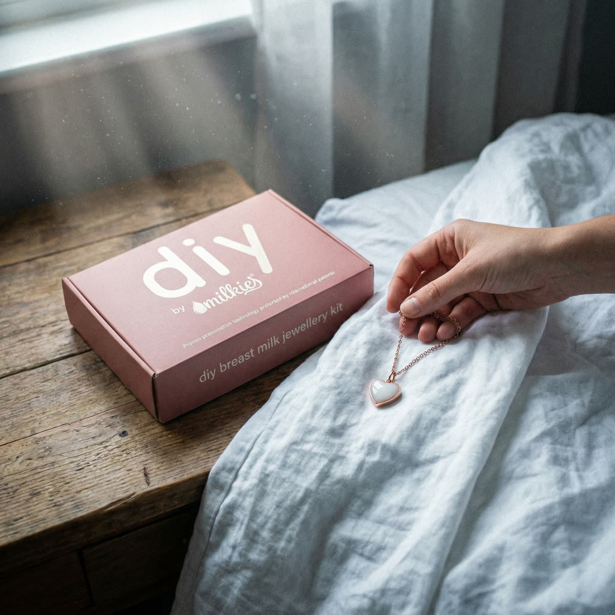 Hand holding a heart-shaped pendant made with an at-home DIY by MILKIES kit for creating breastmilk necklaces, shown beside a pink “DIY breast milk jewellery kit” box on a bedside table.