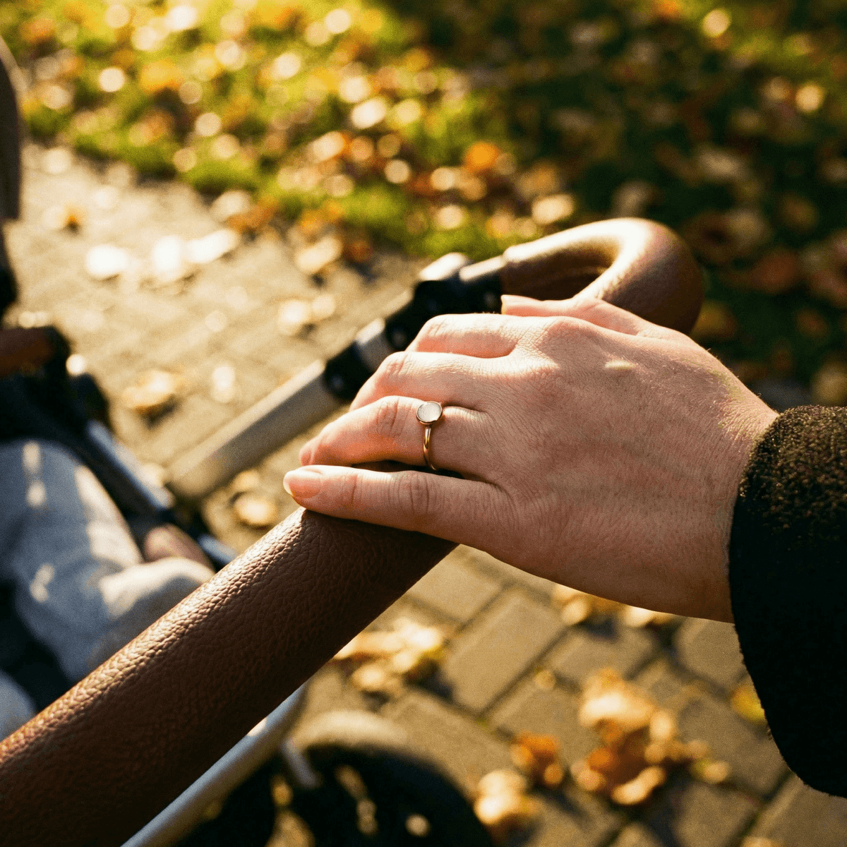 Mother’s hands on a stroller handle outdoors in warm sunlight, wearing a minimalist gold breastmilk ring with a milky white stone