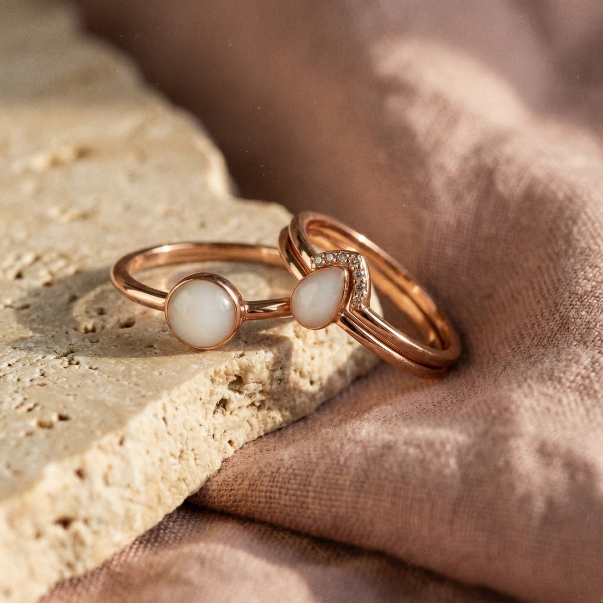Rose gold jewelry set on a natural stone surface featuring a resin breastmilk ring with milky white stones, a matching band, and stud earrings.