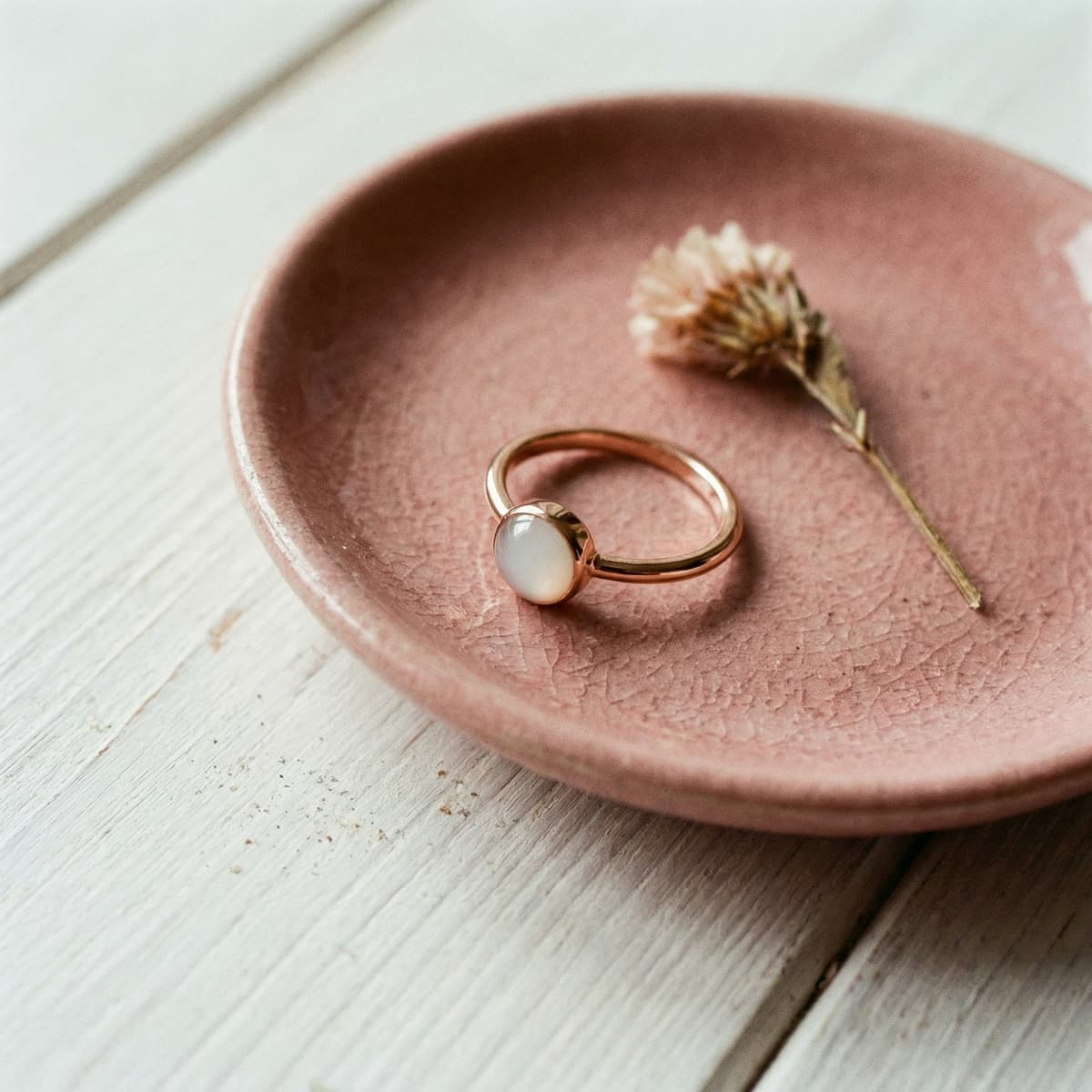 Delicate gold ring with a milky white stone on a pink ceramic dish beside a dried flower, featuring a DIY breastmilk ring kit keepsake design.