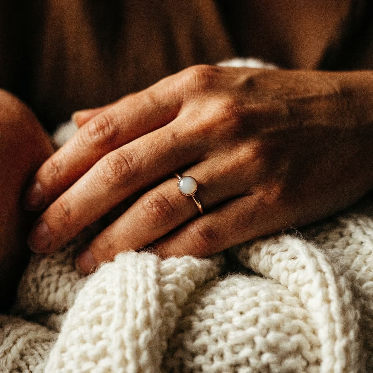 Mother’s hands resting on a knitted blanket, wearing a delicate gold ring with a milky white stone made using a breastmilk ring making kit.