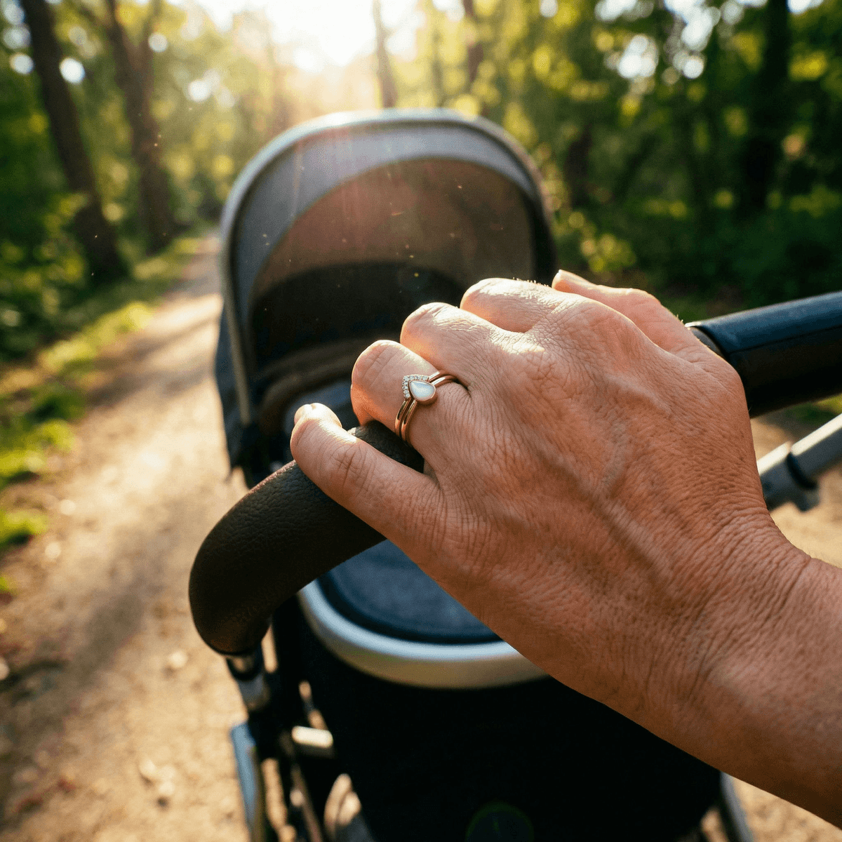 Mother wearing gold breastmilk rings with a milky white teardrop stone while pushing a baby stroller along a sunlit forest path.