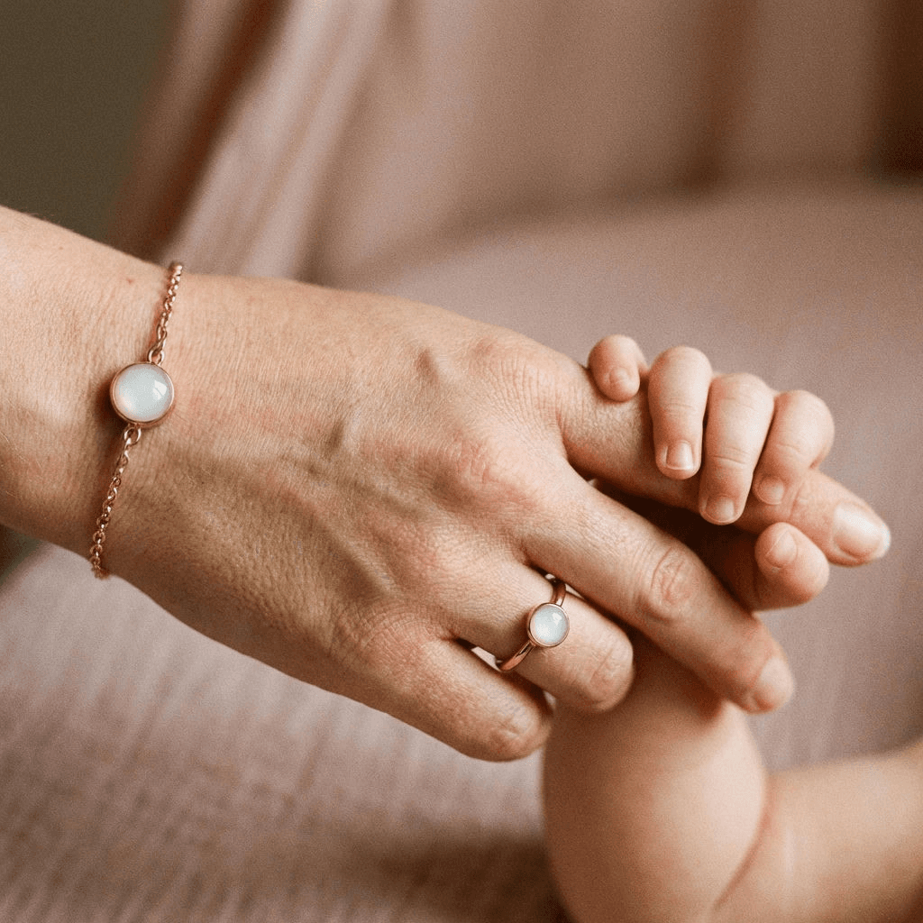 Close-up of a mother holding her baby’s hand, wearing a delicate gold chain with a milky-white stone diy breastmilk bracelet keepsake, paired with a matching ring.
