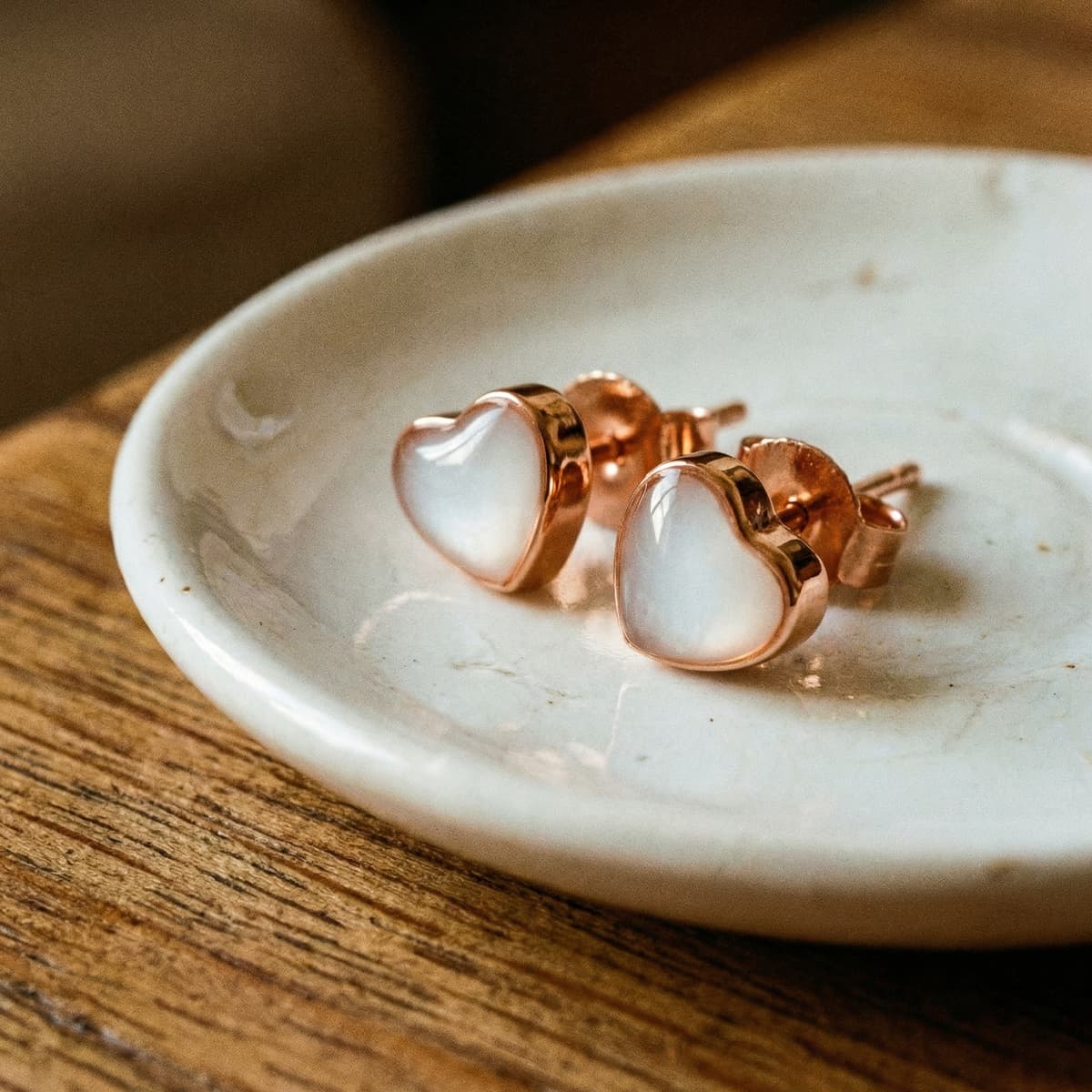 Rose gold heart stud earrings displayed on a ceramic dish with milky white resin centers, showing a diy breastmilk earring keepsake on a wooden surface.