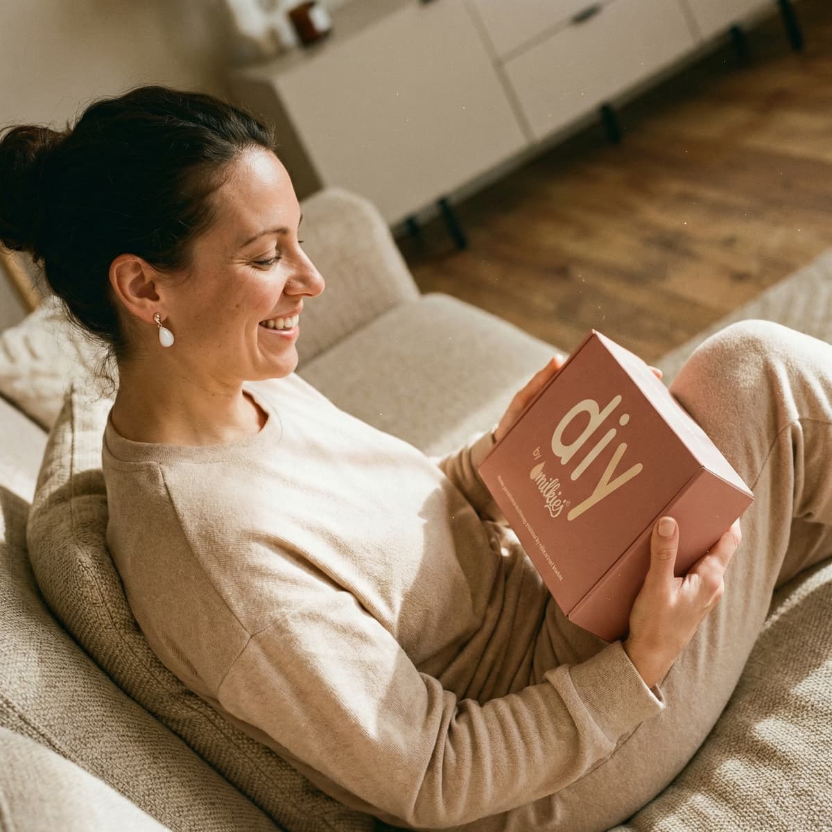 Smiling mother relaxing on a sofa holding a DIY by MILKIES kit box, showing why an at-home diy breastmilk earring keepsake kit is a convenient and private way to create meaningful jewelry.