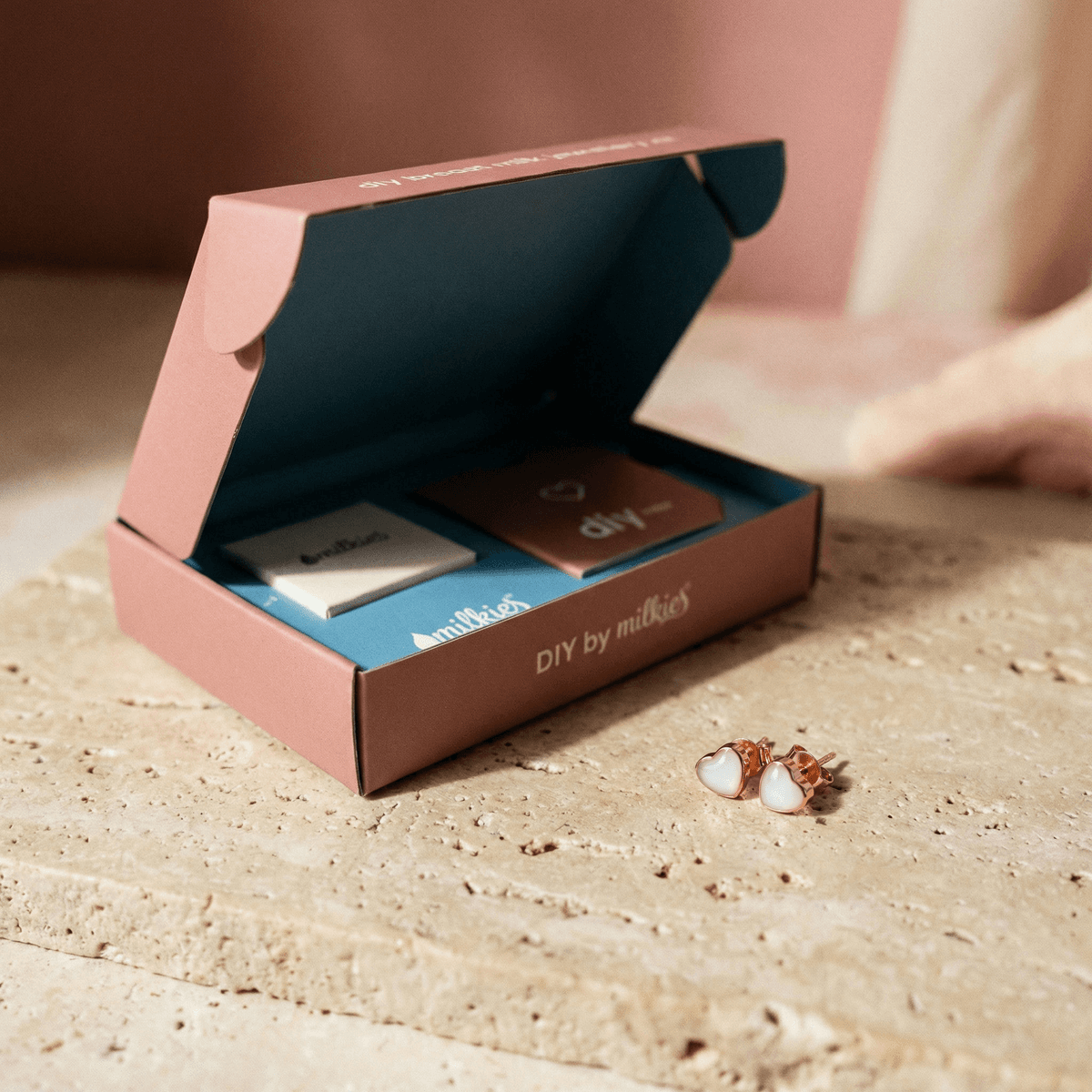 Open DIY by MILKIES at-home keepsake box with tools and pink instruction booklet beside heart-shaped rose-gold stud earrings, showing a diy breastmilk earring kit for preserving breastmilk in resin.