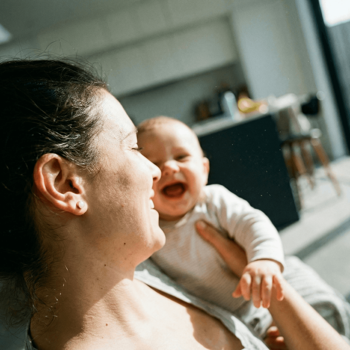 Mother holding a smiling baby in a sunlit home, wearing subtle stud earrings made as a keepsake using a diy breastmilk earring kit for personalized jewelry at home.