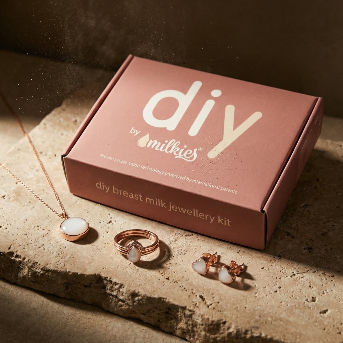Rose-gold DIY by Milkies boxed diy breastmilk jewellery kit on a stone surface, featuring milky white resin pendant necklace, ring, and stud earrings