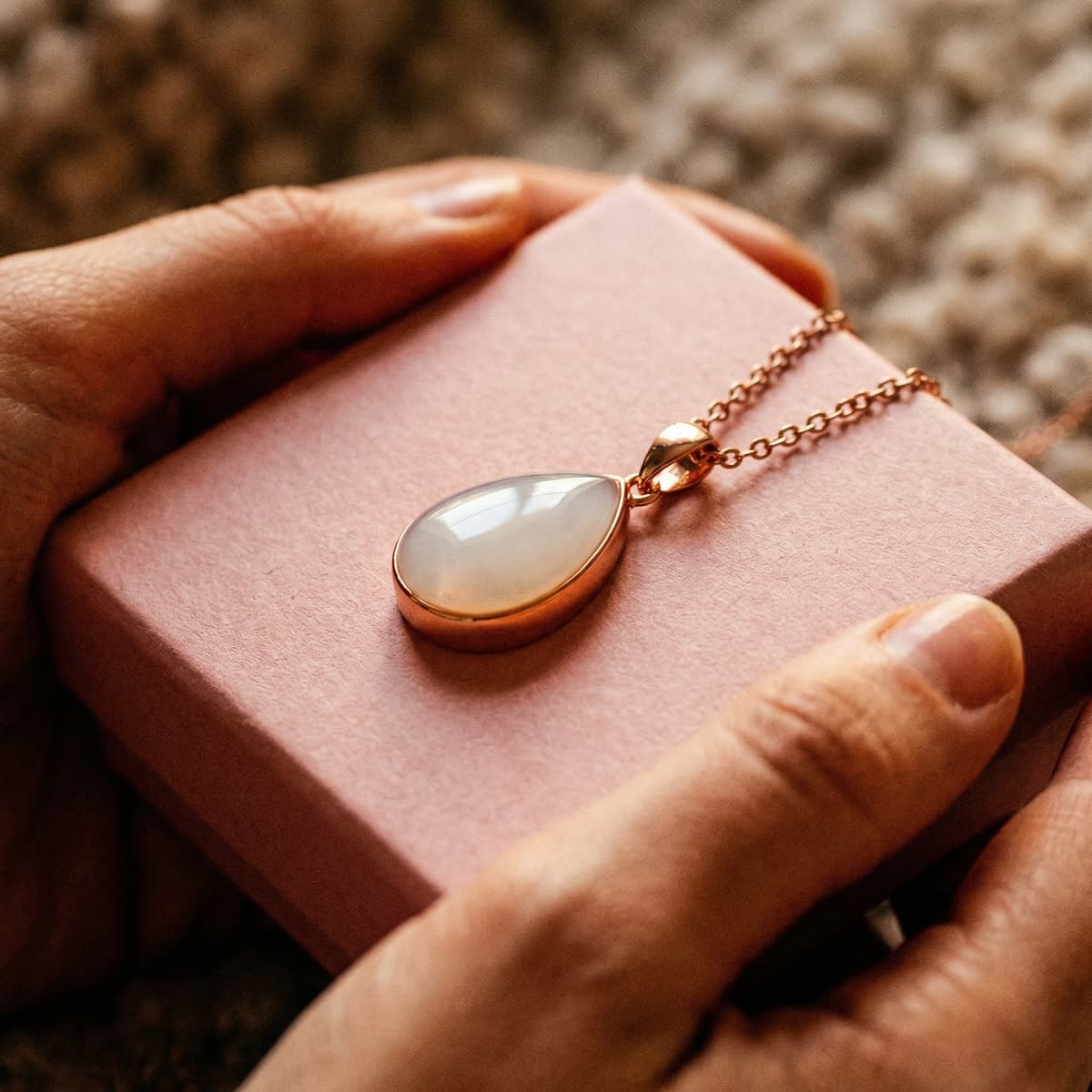 Hands hold a pink gift box with a gold teardrop pendant, showing why a diy breastmilk necklace kit is a meaningful at-home way to preserve breastmilk in jewelry privately.