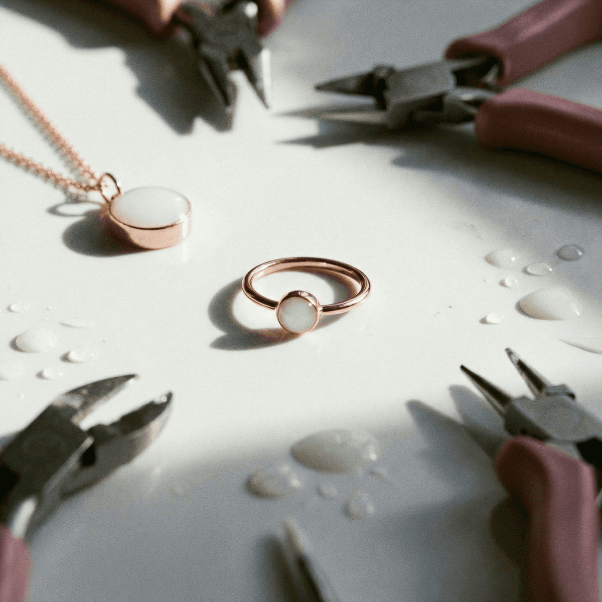Close-up of a rose-gold ring featuring a milky white resin stone made as a diy breastmilk ring on a work surface, surrounded by jewelry pliers, a matching pendant, and small drops of liquid.