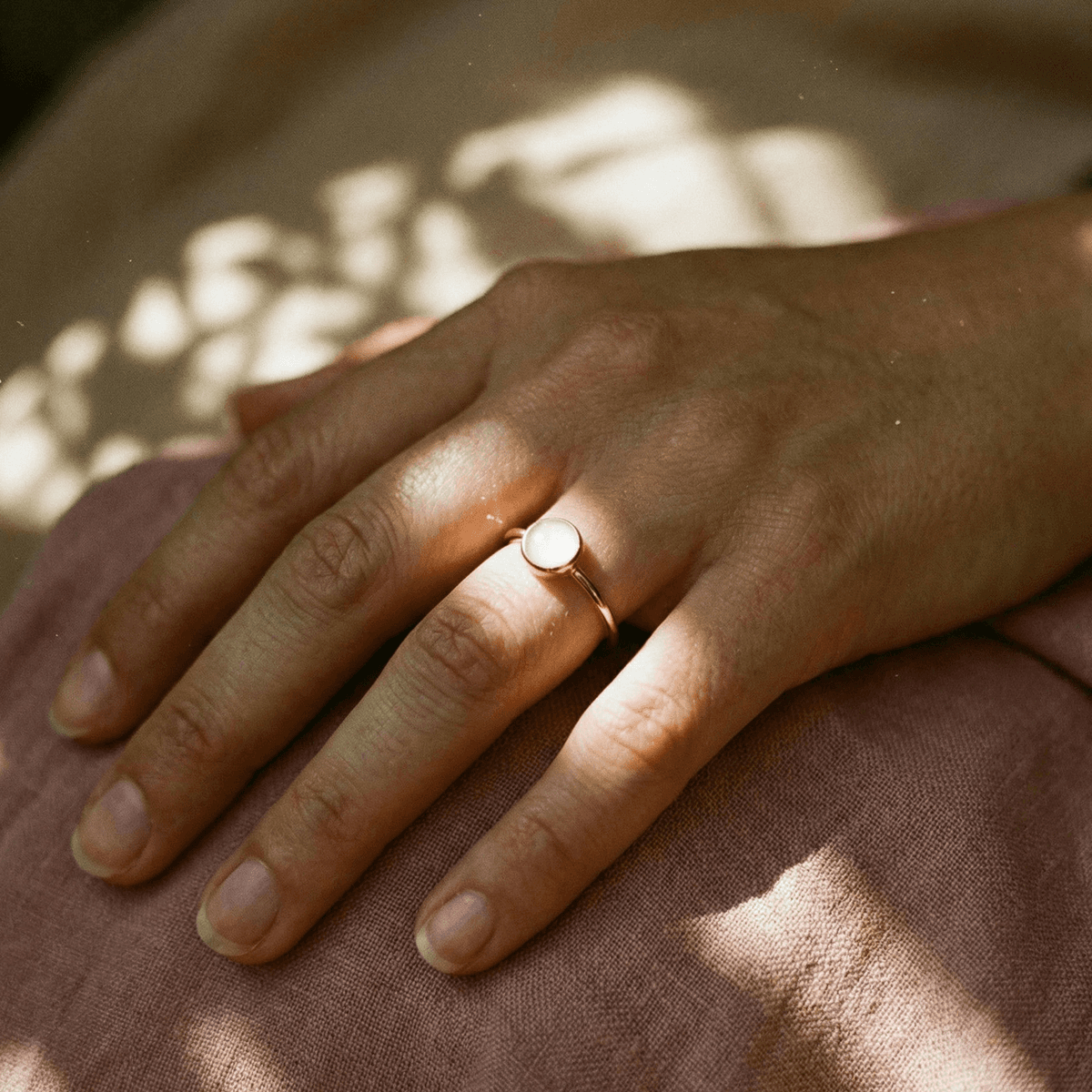 Minimalist gold band ring with a round milky-white stone on a hand, showcasing a diy breastmilk ring in warm natural light against soft pink fabric.