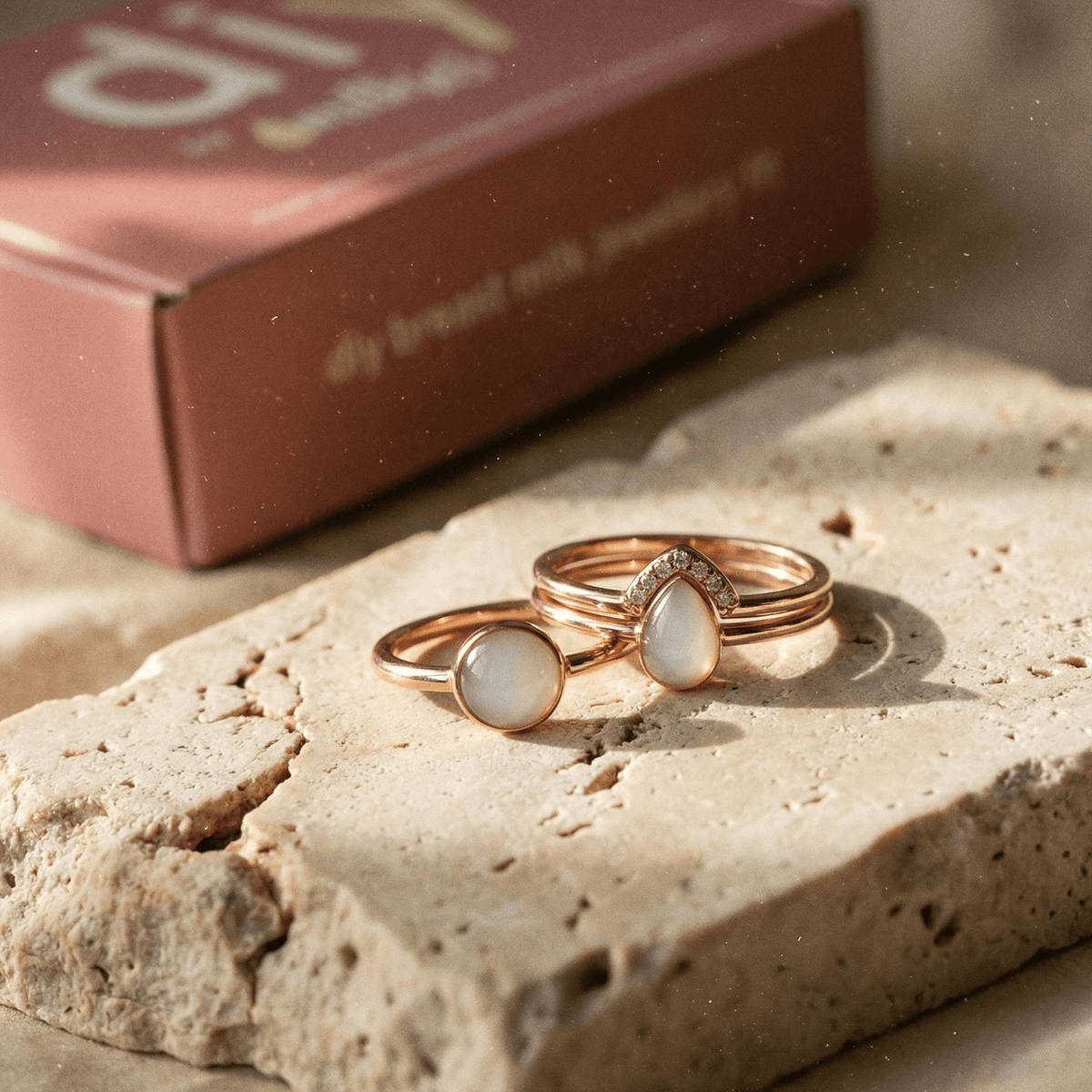 Rose gold keepsake rings with milky white resin stones on a natural stone slab next to a branded box, representing a diy breastmilk ring kit for making breastmilk jewelry at home.