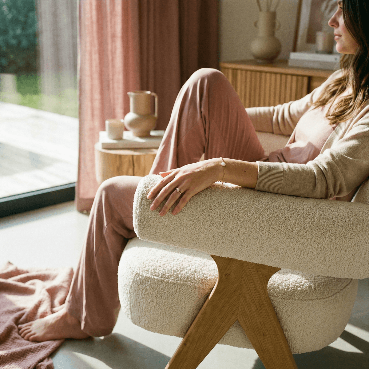 Woman in pink loungewear relaxing in a cozy armchair by a sunlit window, wearing delicate jewelry and practicing calm self-care while drying up milk.
