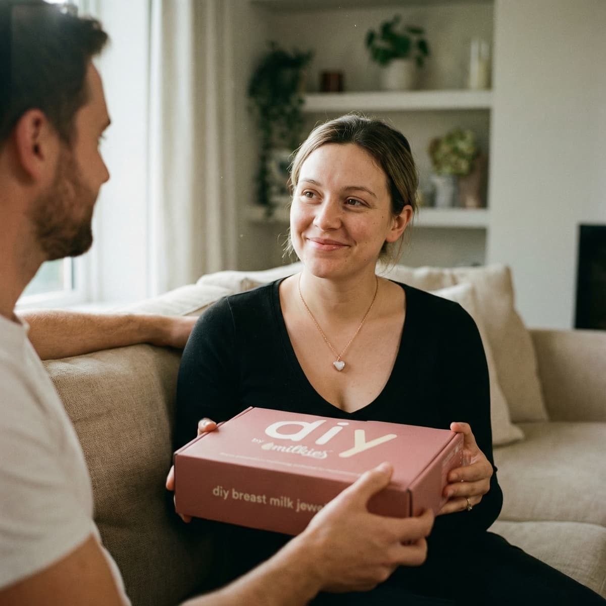 Woman smiling on a sofa receiving a pink DIY by MILKIES at-home breastmilk keepsake jewelry kit box, a meaningful gift for new mother and a hands-on way to preserve precious memories.