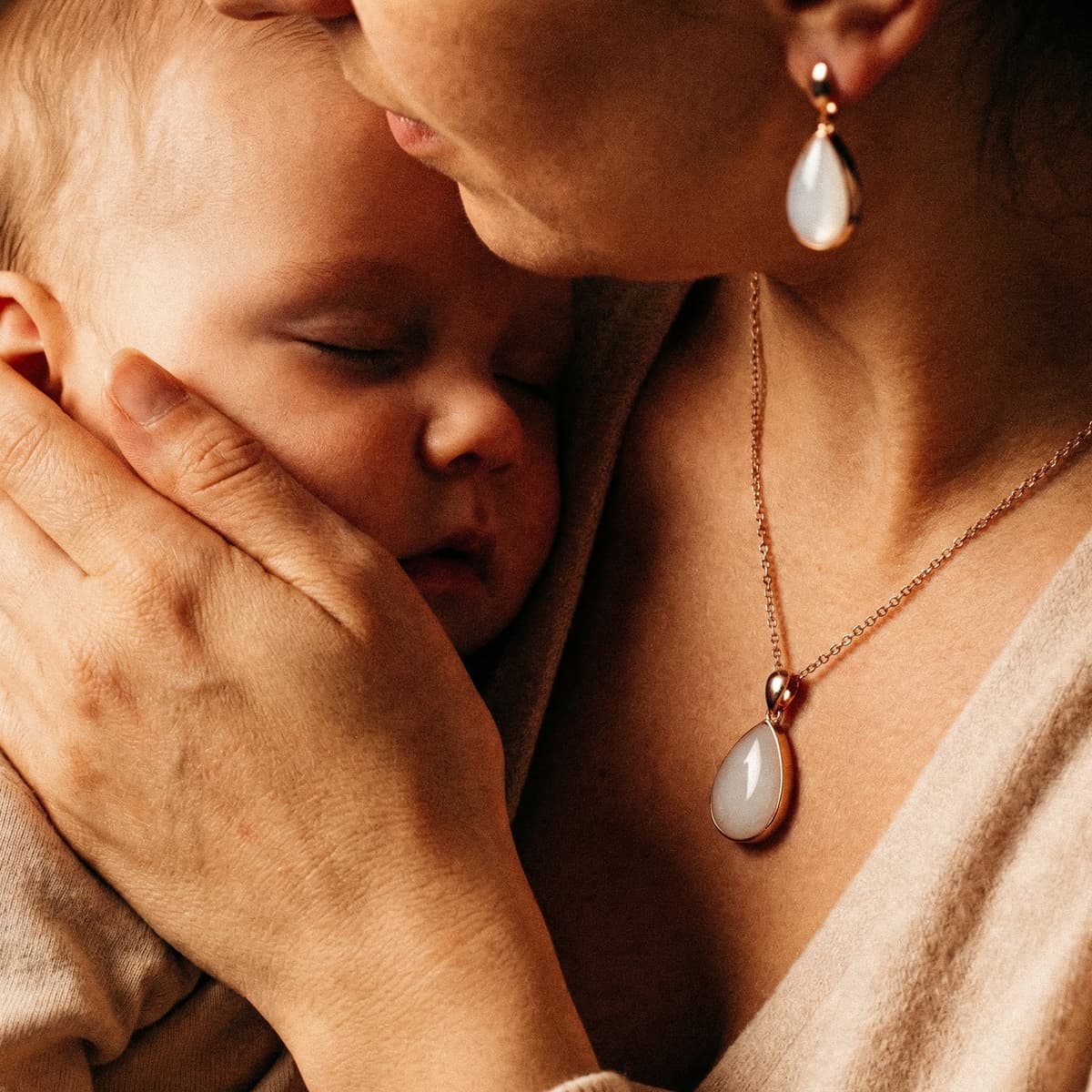 Mother cuddling a sleeping newborn while wearing elegant breastmilk keepsake jewelry, including a teardrop white stone pendant necklace and matching earrings in gold—golden boobs close-up.