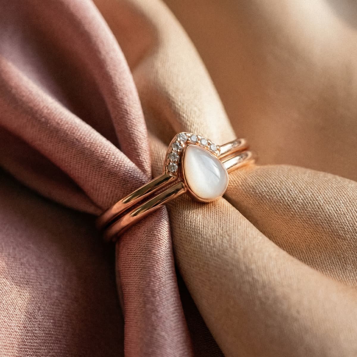 Rose gold ring featuring a teardrop-shaped milky white stone with small crystal accents, photographed on blush and beige fabric as a delicate motherhood keepsake jewelry piece.