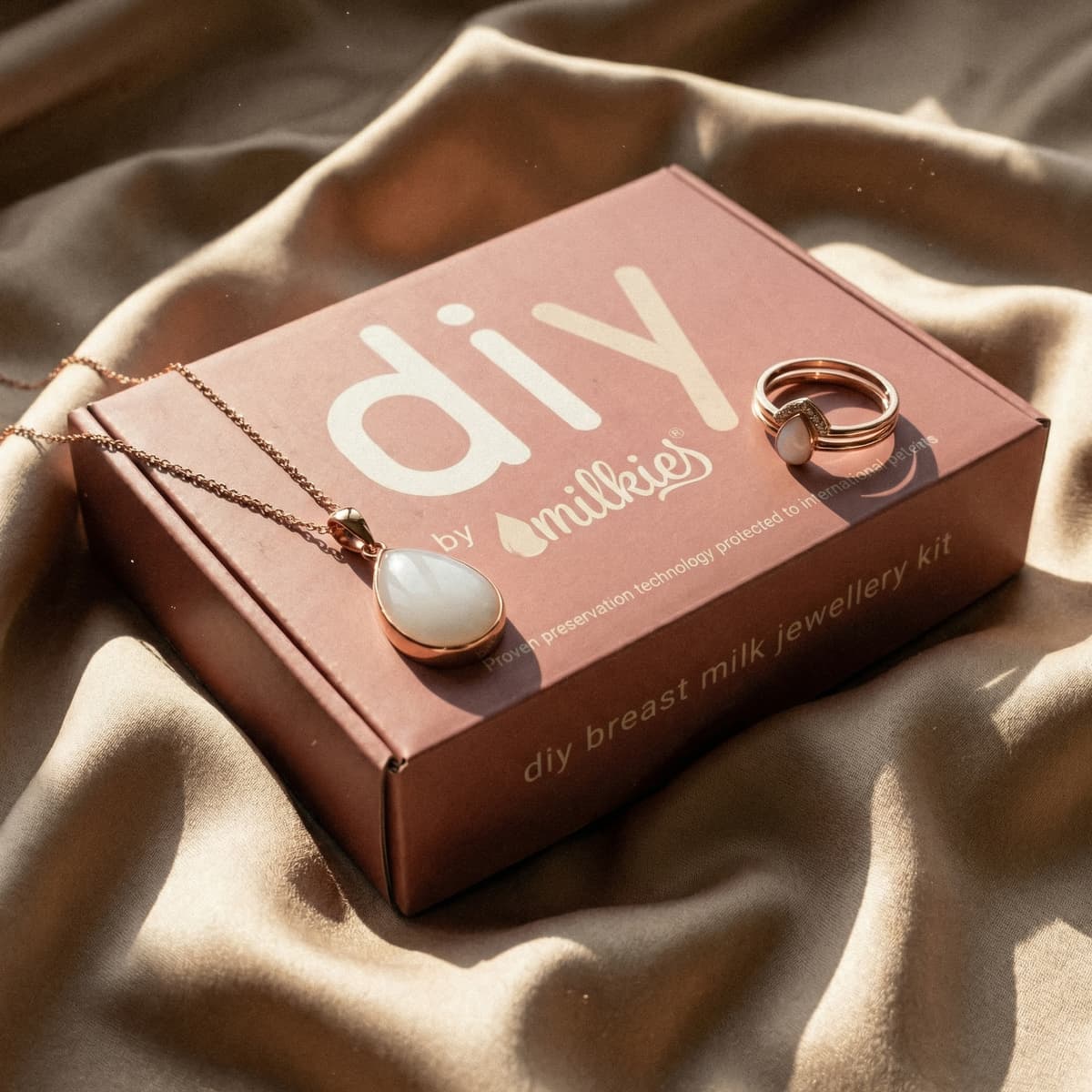 DIY by MILKIES breast milk jewelry kit in a pink box on soft fabric, a thoughtful option for new mom gifts, featuring a rose-gold teardrop milk-stone necklace and matching ring.